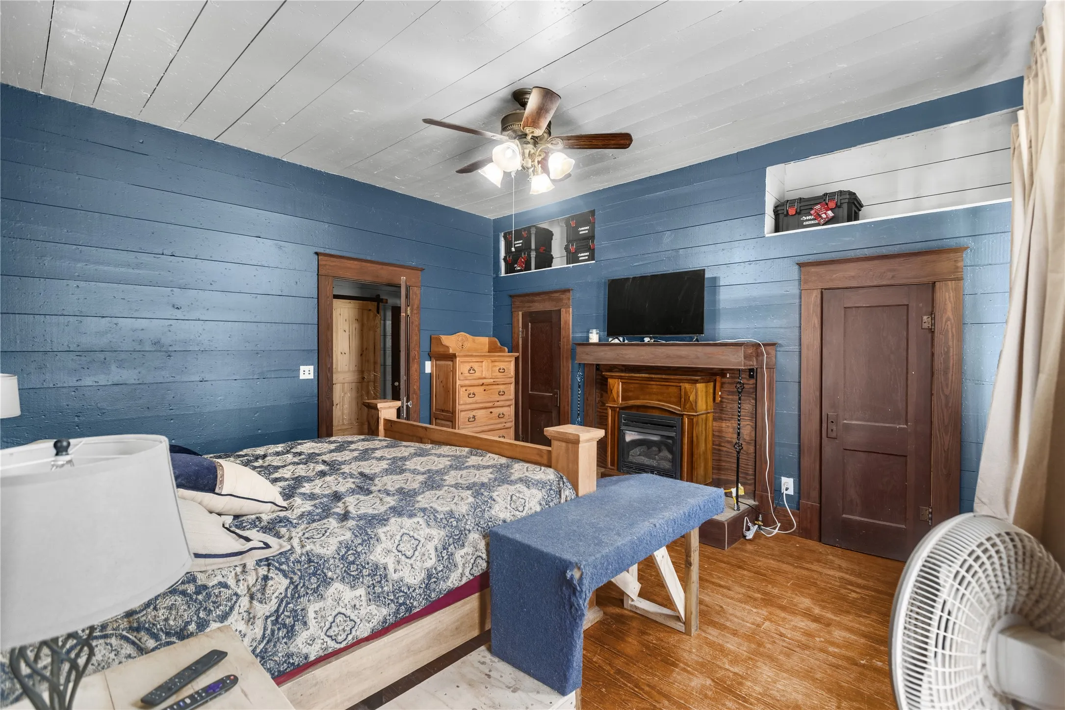 Spacious bedroom featuring shiplap walls in a rich blue finish, hardwood flooring, ceiling fan with integrated lighting, built-in wooden dresser, and a fireplace insert with wooden mantel