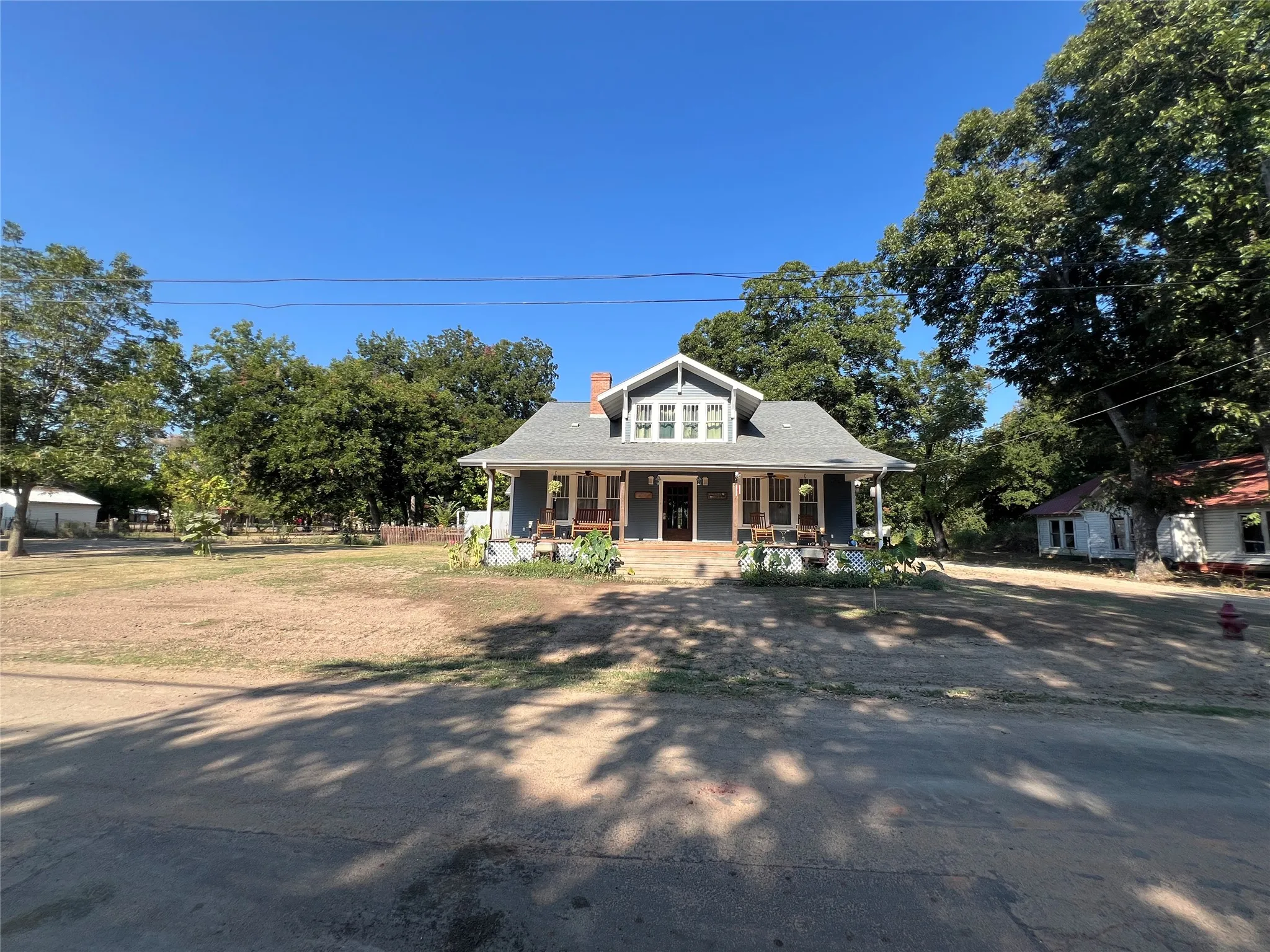 Gabled roofline with dormer window, covered porch with columns, brick chimney, dark siding, and mature trees
