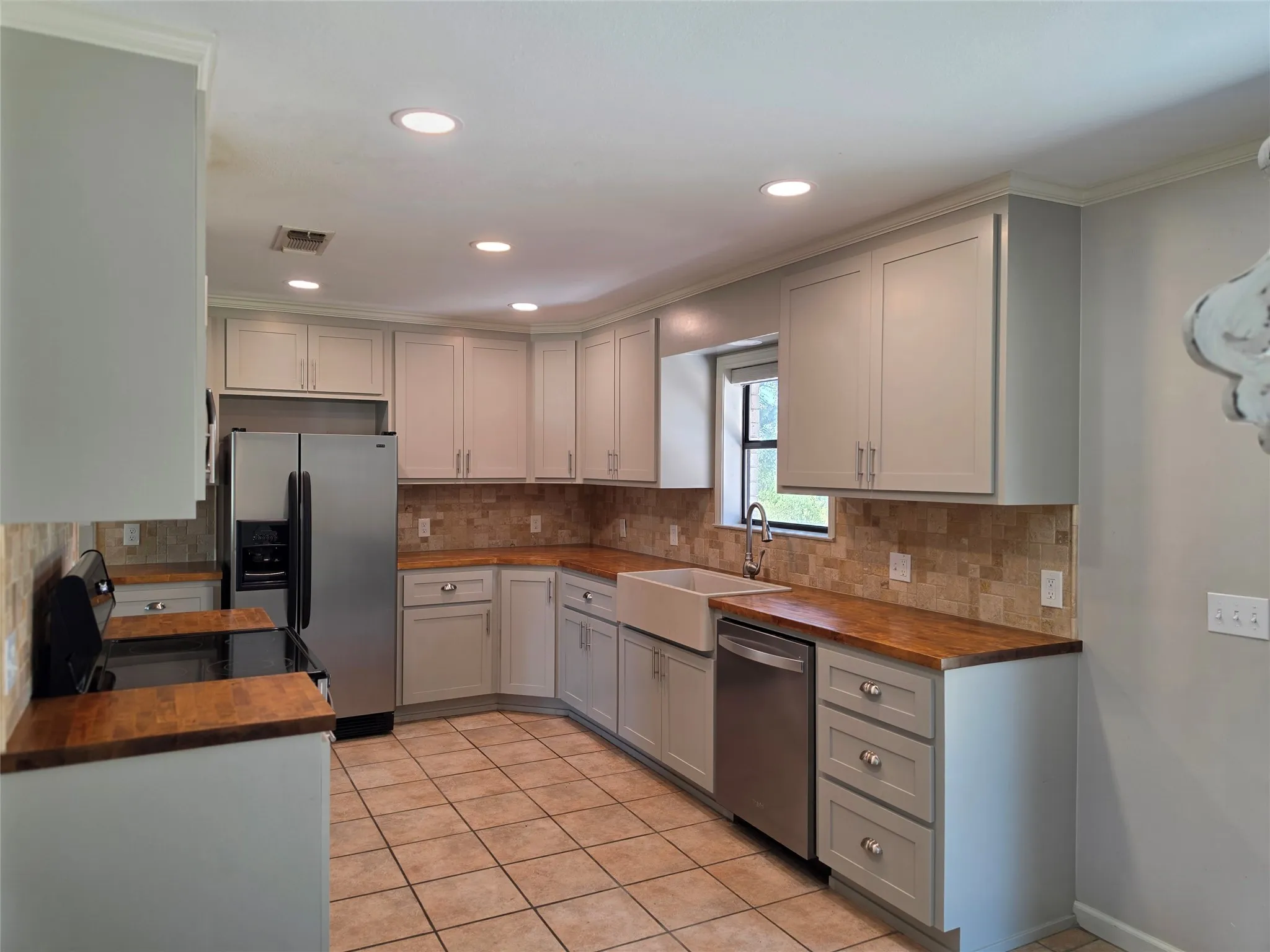 Recessed lighting illuminates the kitchen featuring wood-finish countertops, light-toned cabinetry, a farmhouse-style sink, and stainless steel appliances