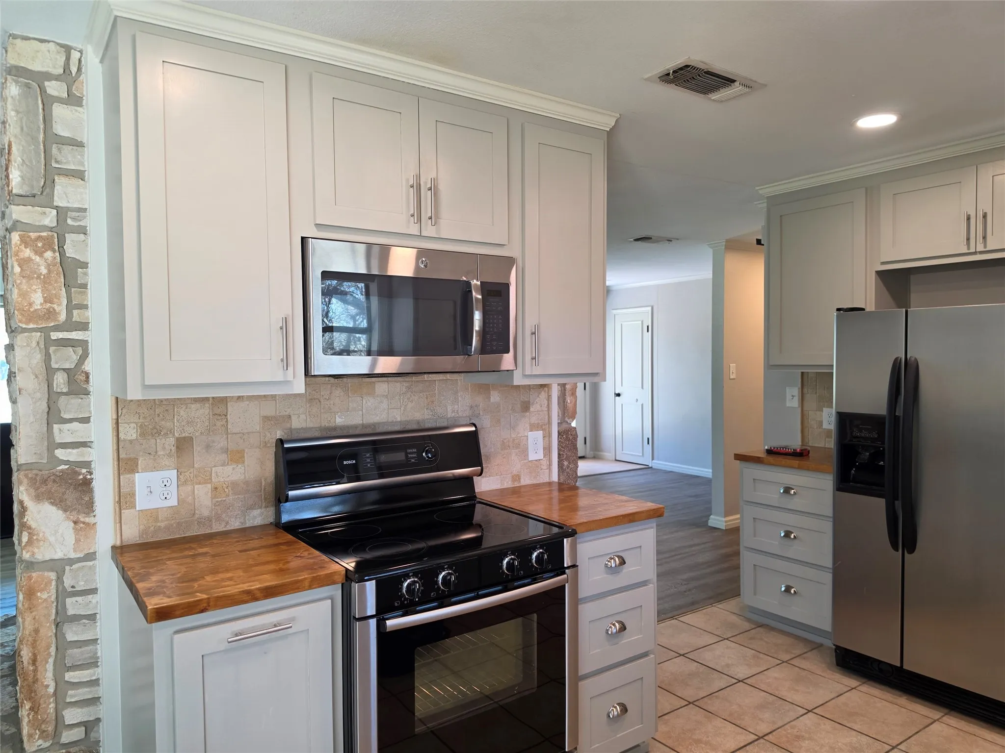 Shaker-style cabinetry with crown molding, stainless steel appliances, stone tile backsplash, wood-finish countertops, and tile flooring