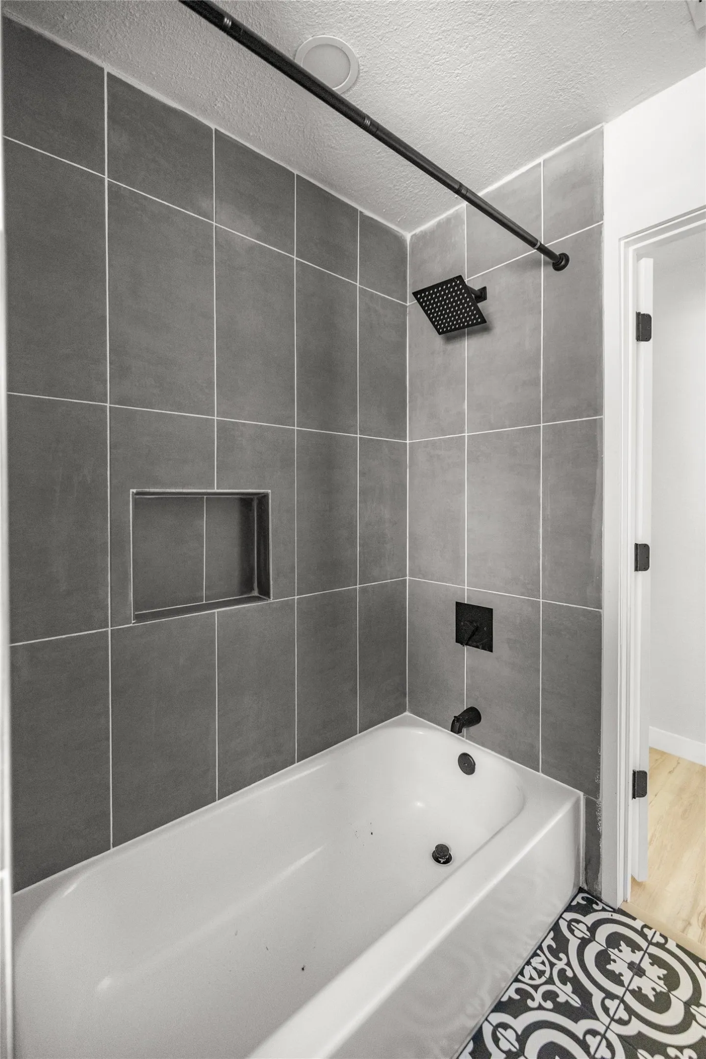 Bathtub with gray tile surround, recessed shelf, matte black shower fixtures, and decorative floor tiling