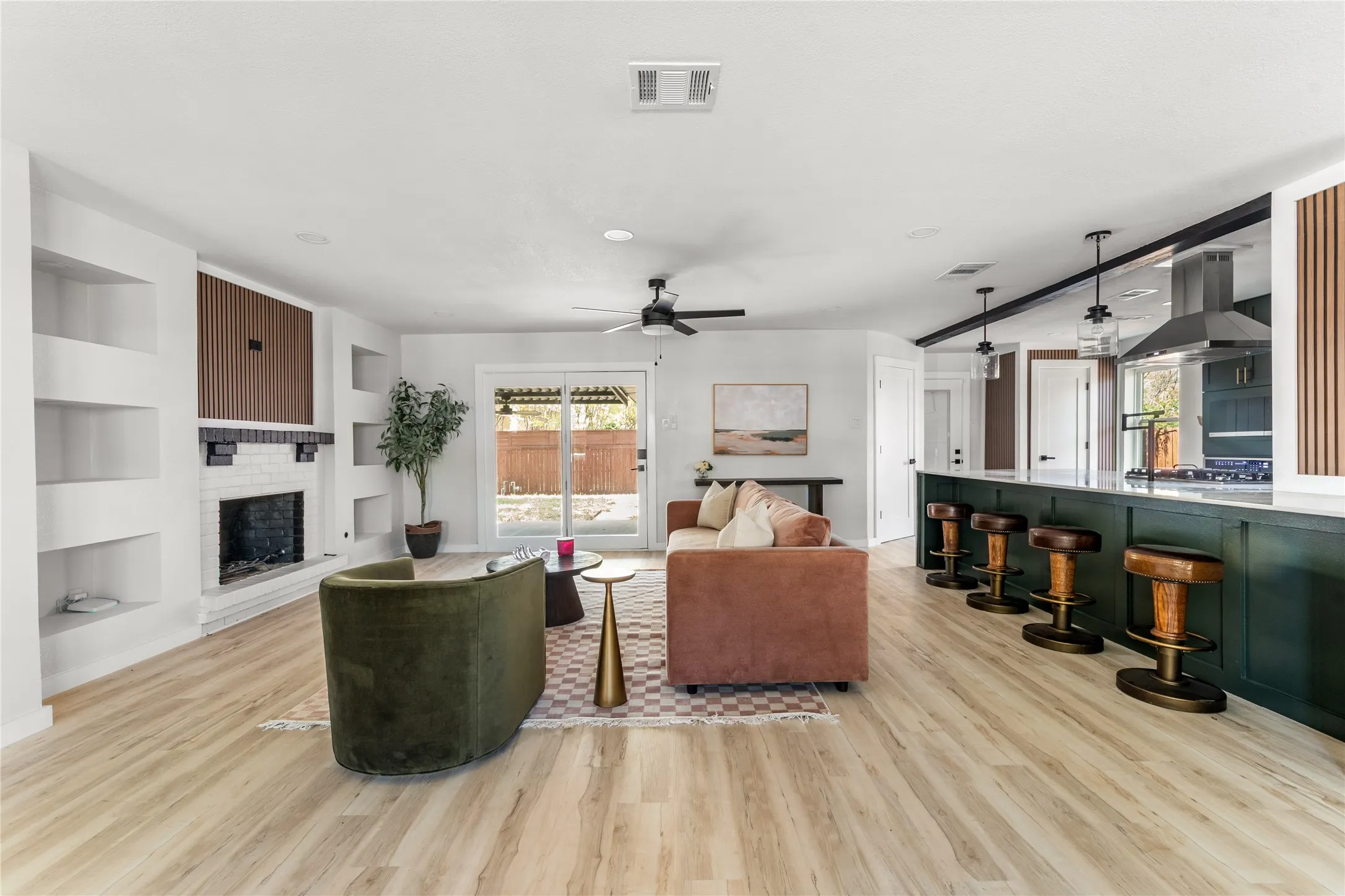 Light wood-finish flooring, built-in shelving, white brick fireplace, dark green paneled kitchen island, and a stainless steel range hood