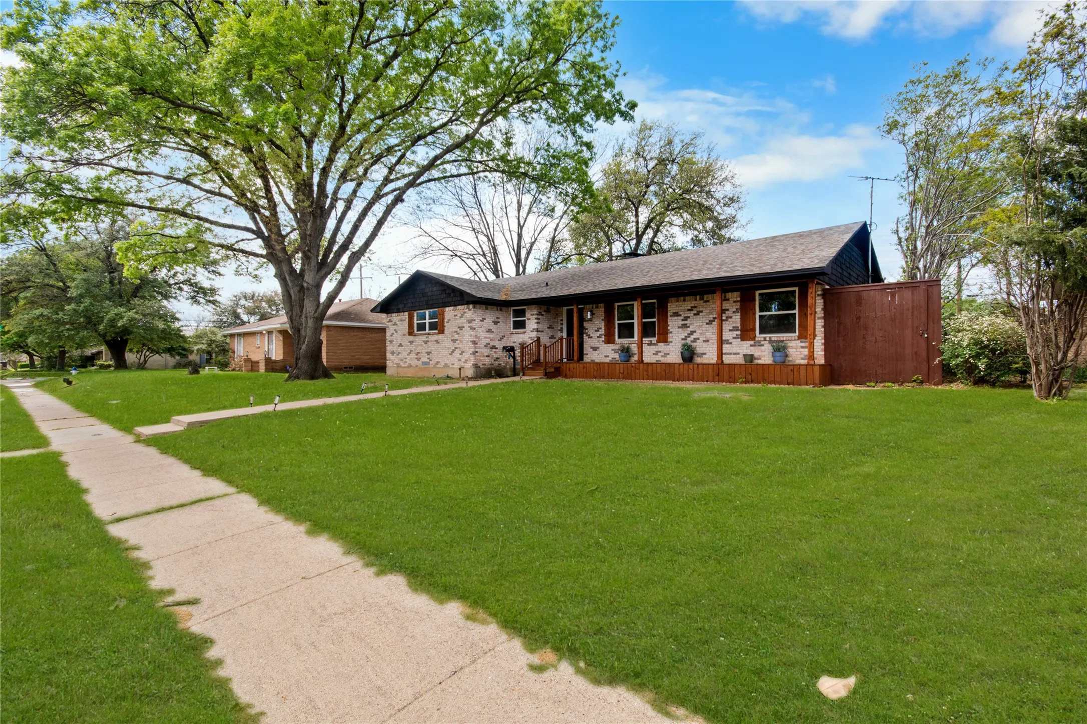 Brick exterior with dark wood accents, a brown shingle roof, a front wood deck, and a sidewalk entrance
