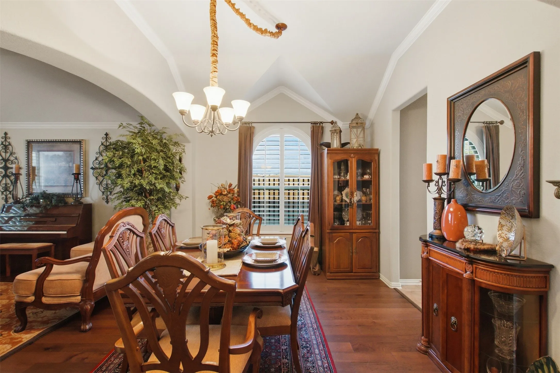 Vaulted ceiling with crown molding, wood-finish flooring, arched entryway, window with plantation shutters, and a built-in display cabinet