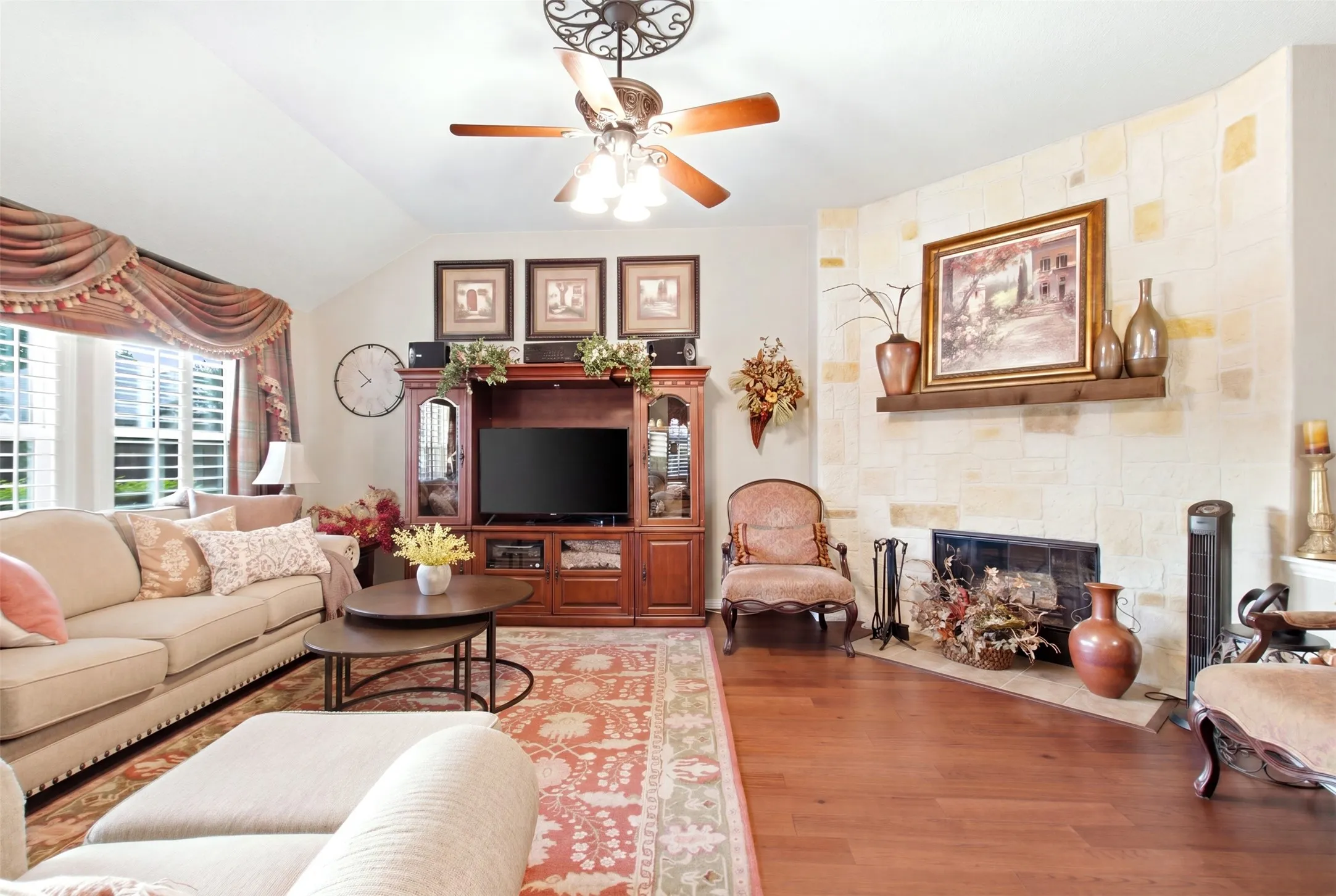 Vaulted ceiling with a ceiling fan, wood-finish flooring, built-in entertainment center, and a stone-clad fireplace with a wood mantel
