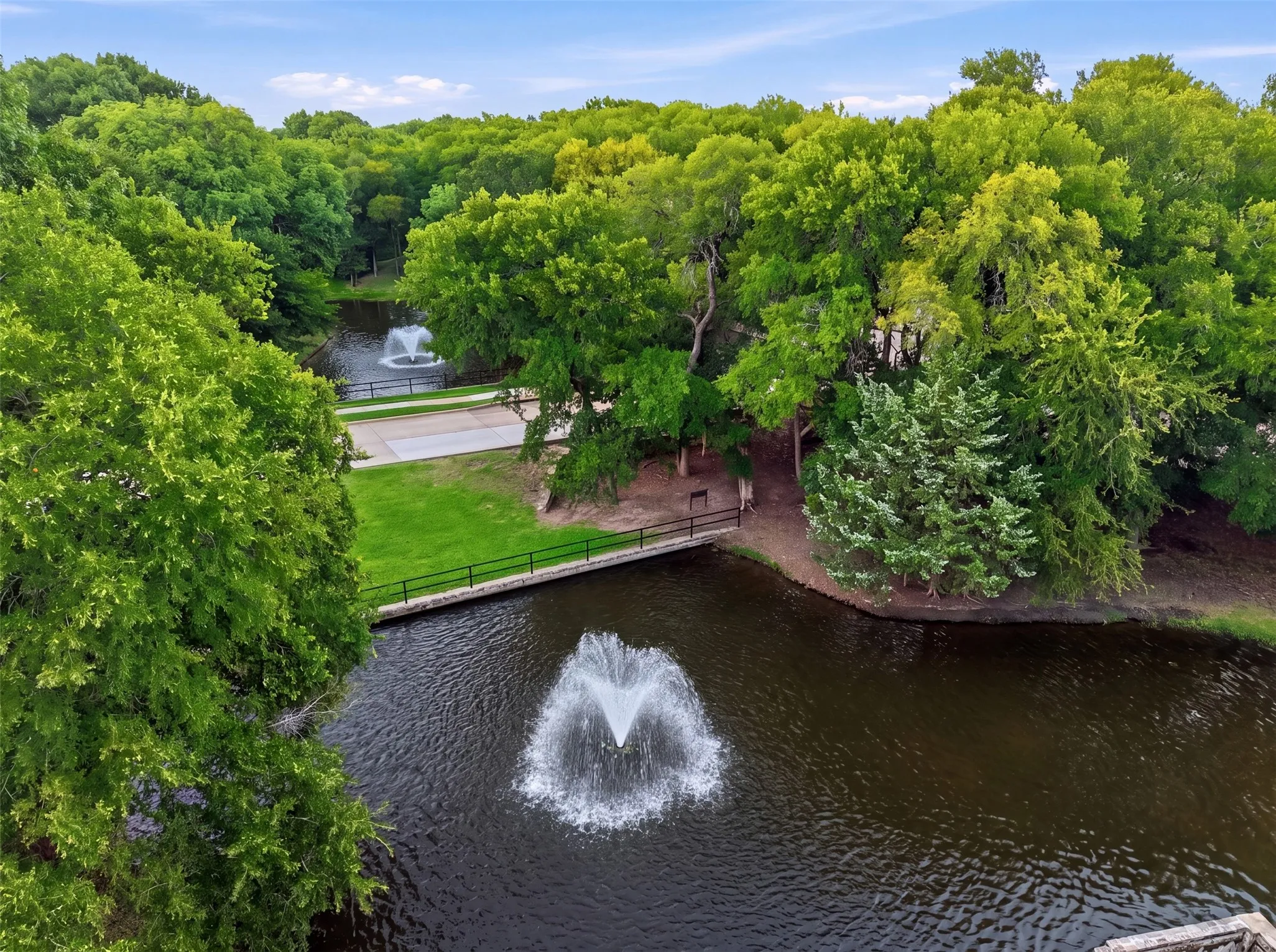 Water features with fountains, surrounding mature trees, manicured grass areas, and a paved pathway