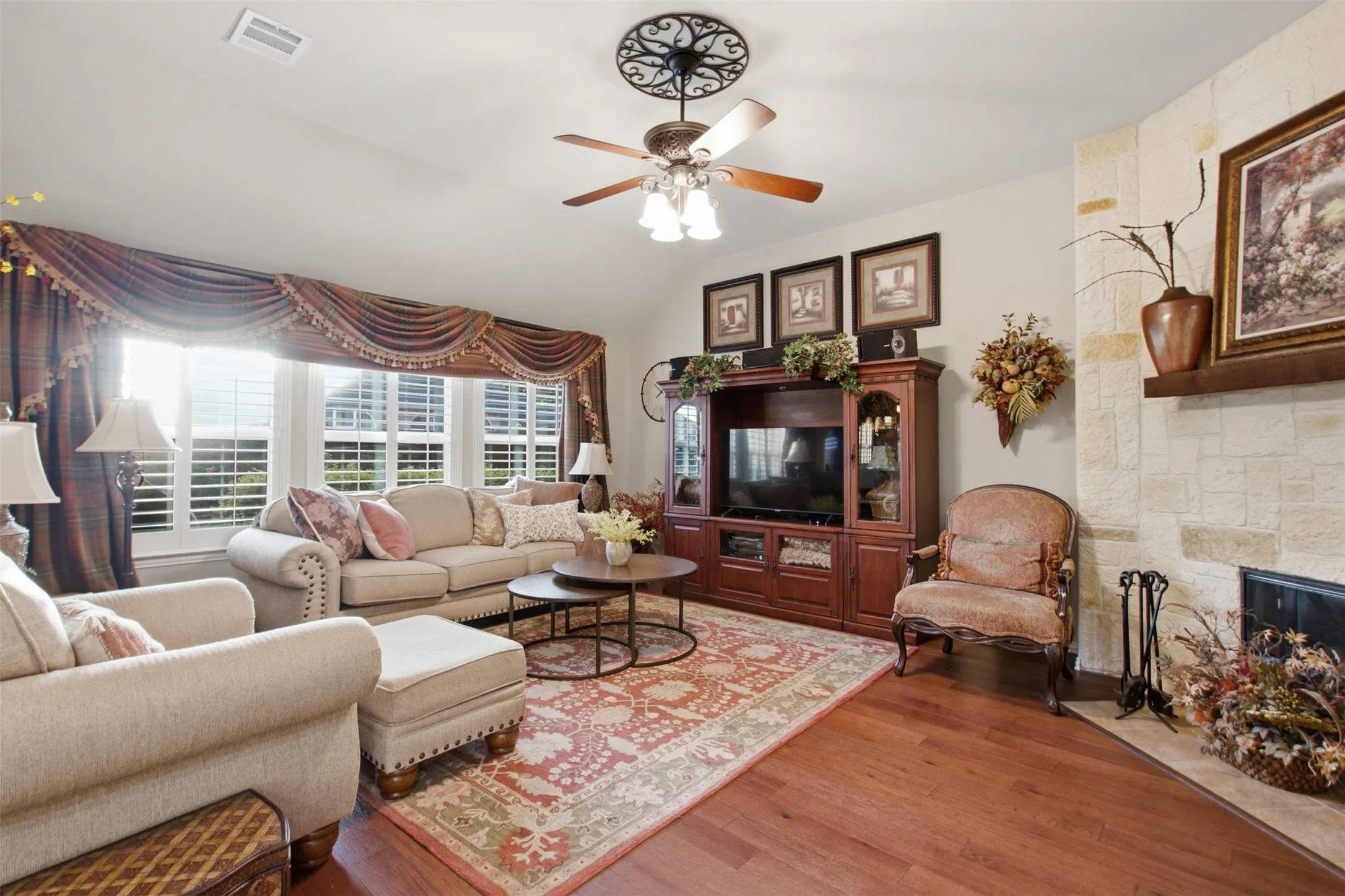 Hardwood-finish flooring, stone-clad fireplace with mantel, window with plantation shutters, ceiling fan with ornate medallion, built-in entertainment cabinetry