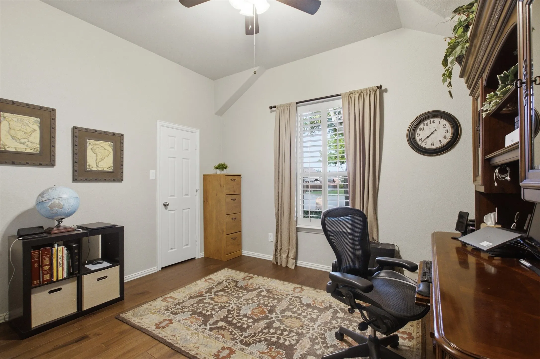 Vaulted ceiling with a ceiling fan, wood-finish flooring, window with plantation shutters, interior door, and built-in shelving unit