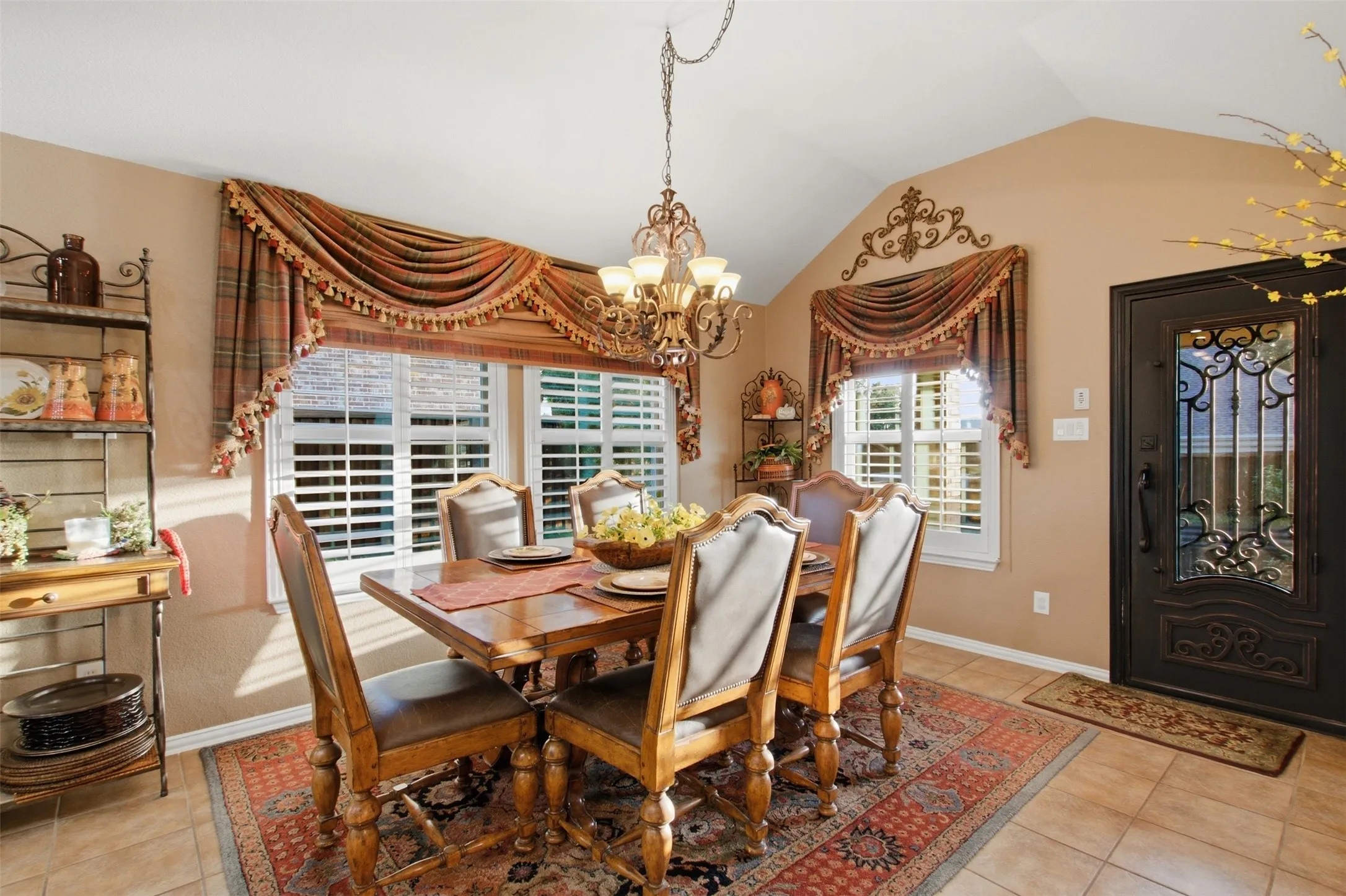 Vaulted ceiling design, decorative chandelier, transom window, plantation shutters, and an ornate front door with glass and wrought iron detailing