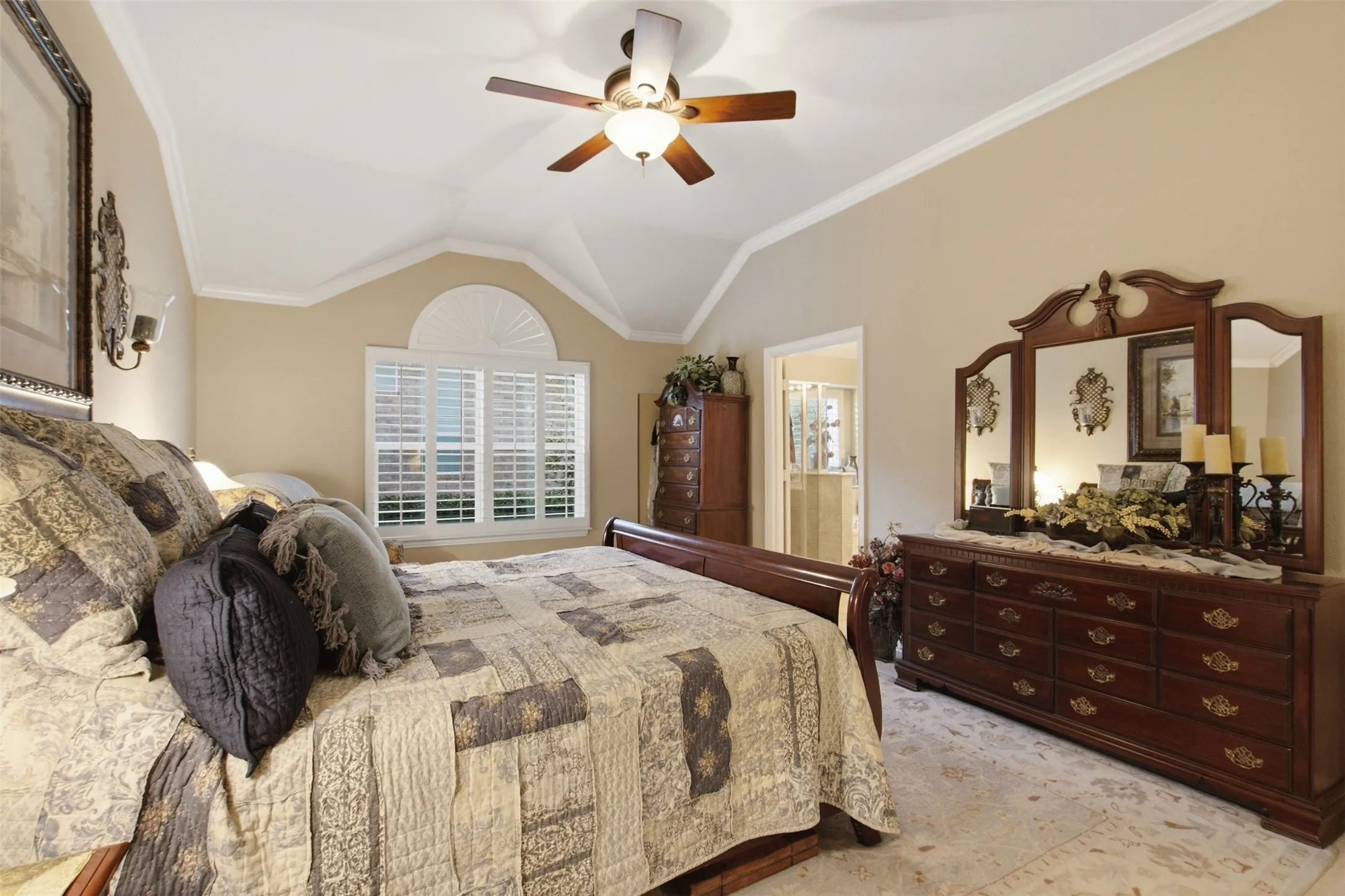 Vaulted ceiling with crown molding, ceiling fan, arched window with plantation shutters, sconce lighting, and a doorway opening to an additional room