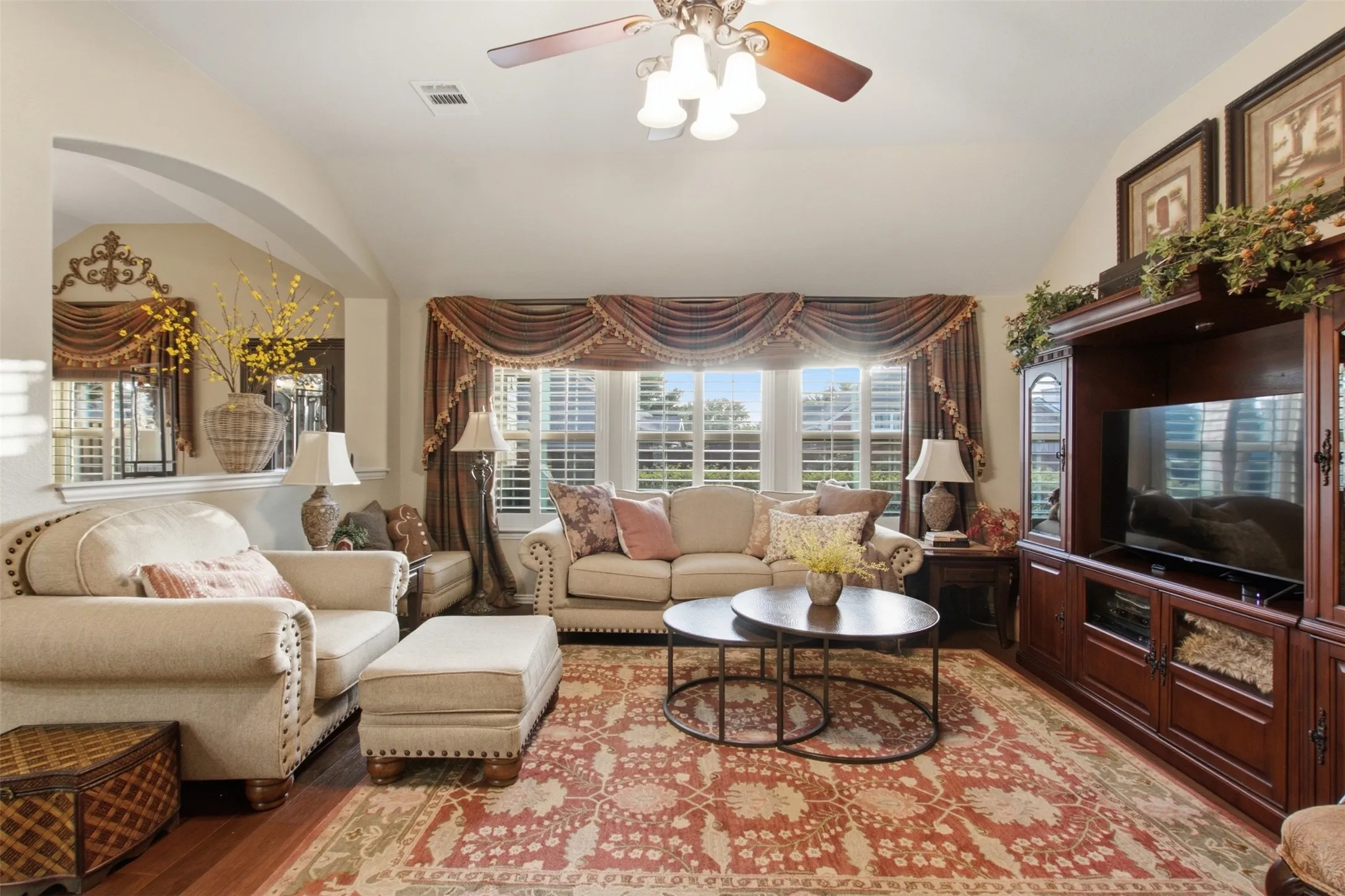 Vaulted ceiling with an attached ceiling fan, bay window with plantation shutters, wood-finish flooring, and a built-in media cabinet