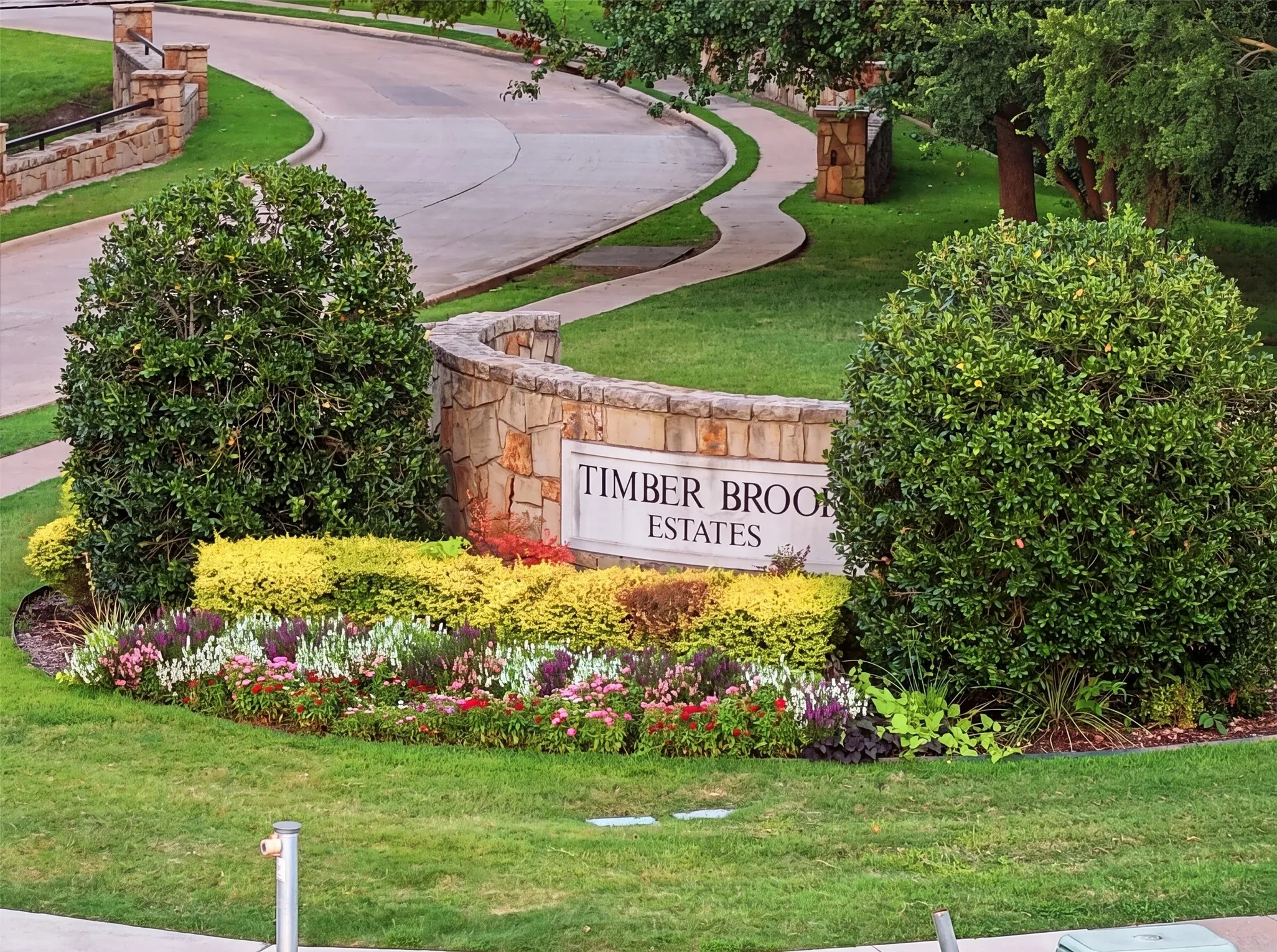 Stone entry monument with engraved signage, curved concrete sidewalk, lush green lawn, and a vibrant flower bed with varied plantings