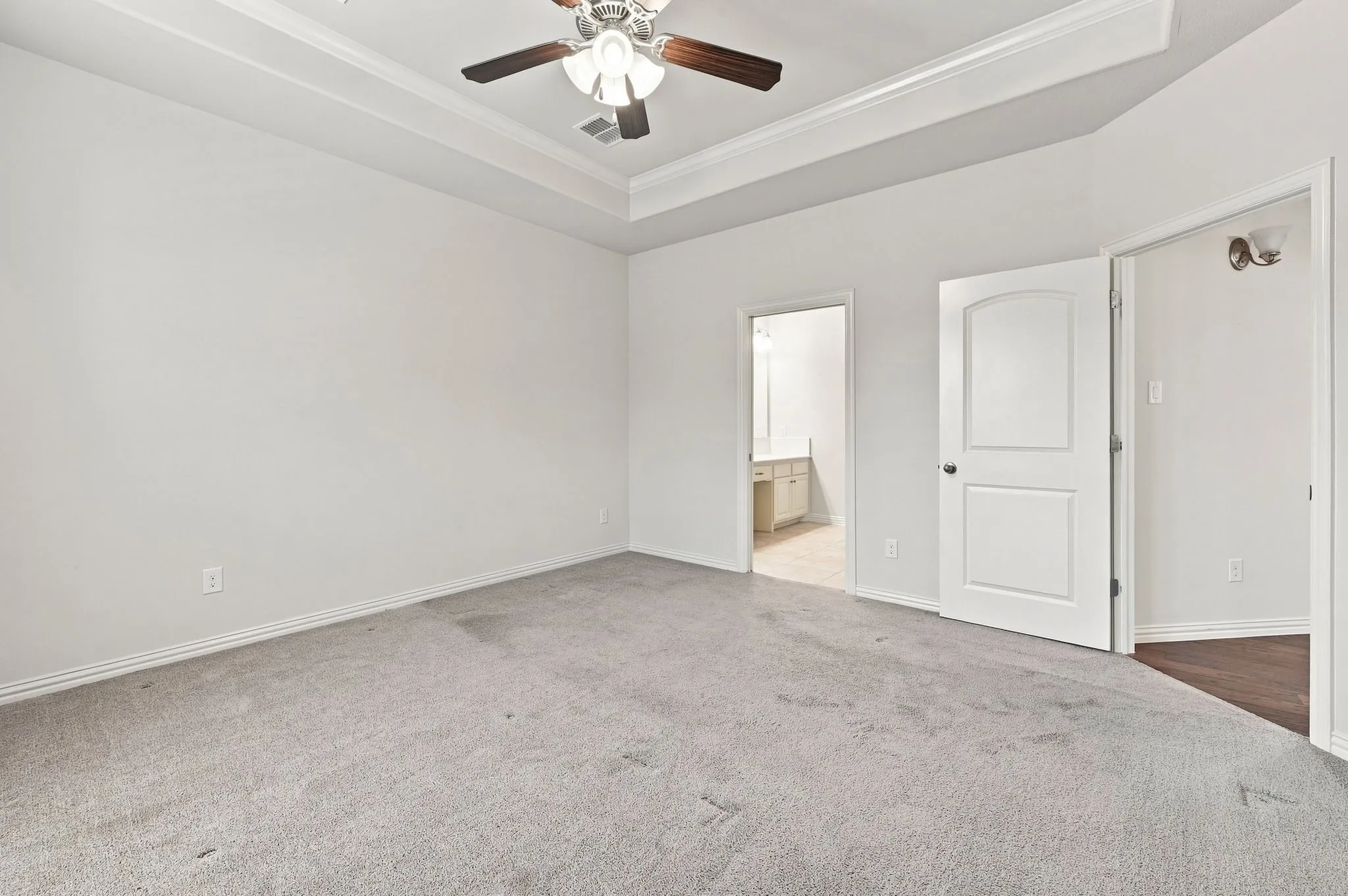 Recessed ceiling with crown molding, ceiling fan with light fixture, light gray carpeting, white paneled doors, and a vanity visible through the doorway