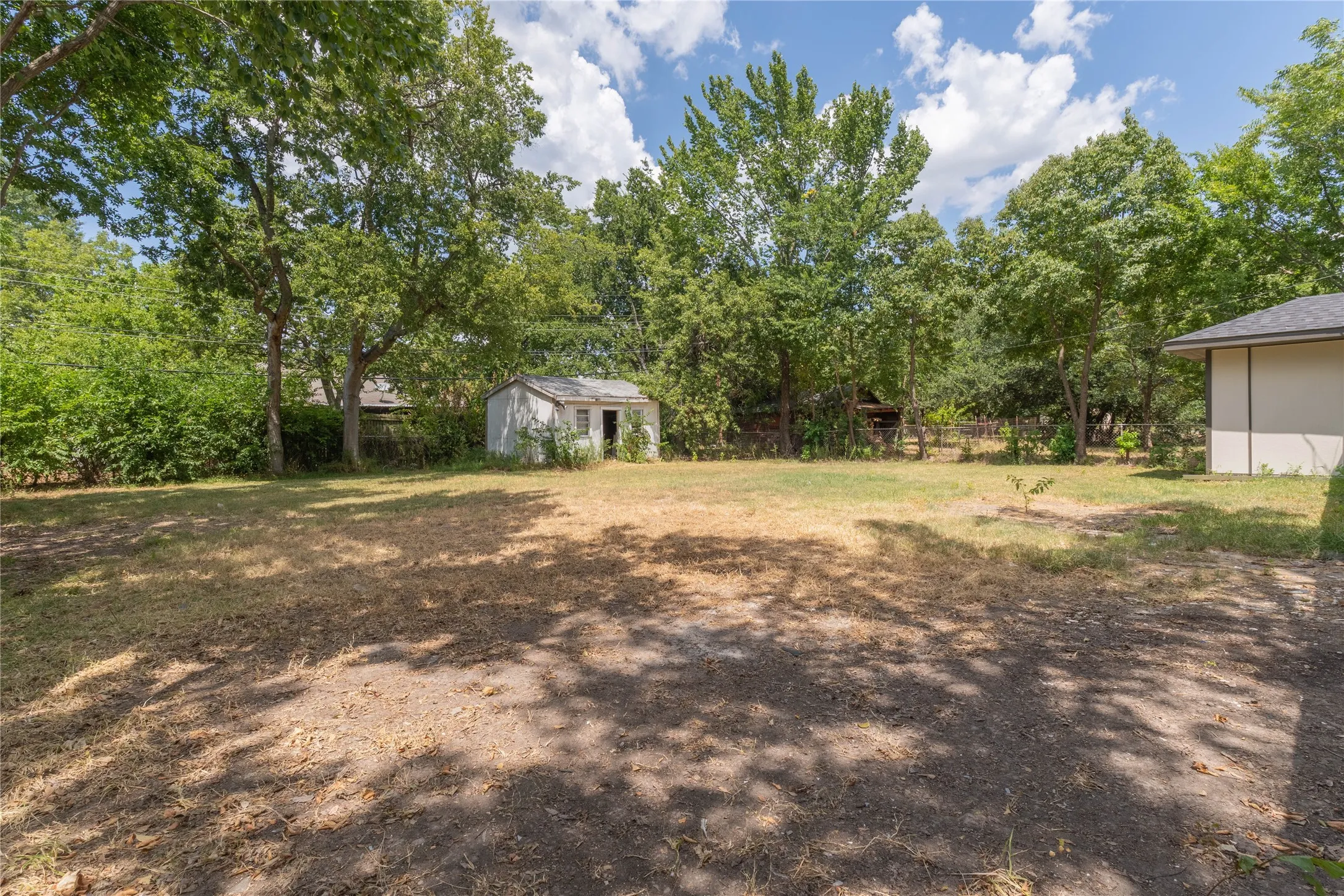 Expansive yard with mature trees providing ample shade, featuring an exterior storage shed and a visible side elevation of the property