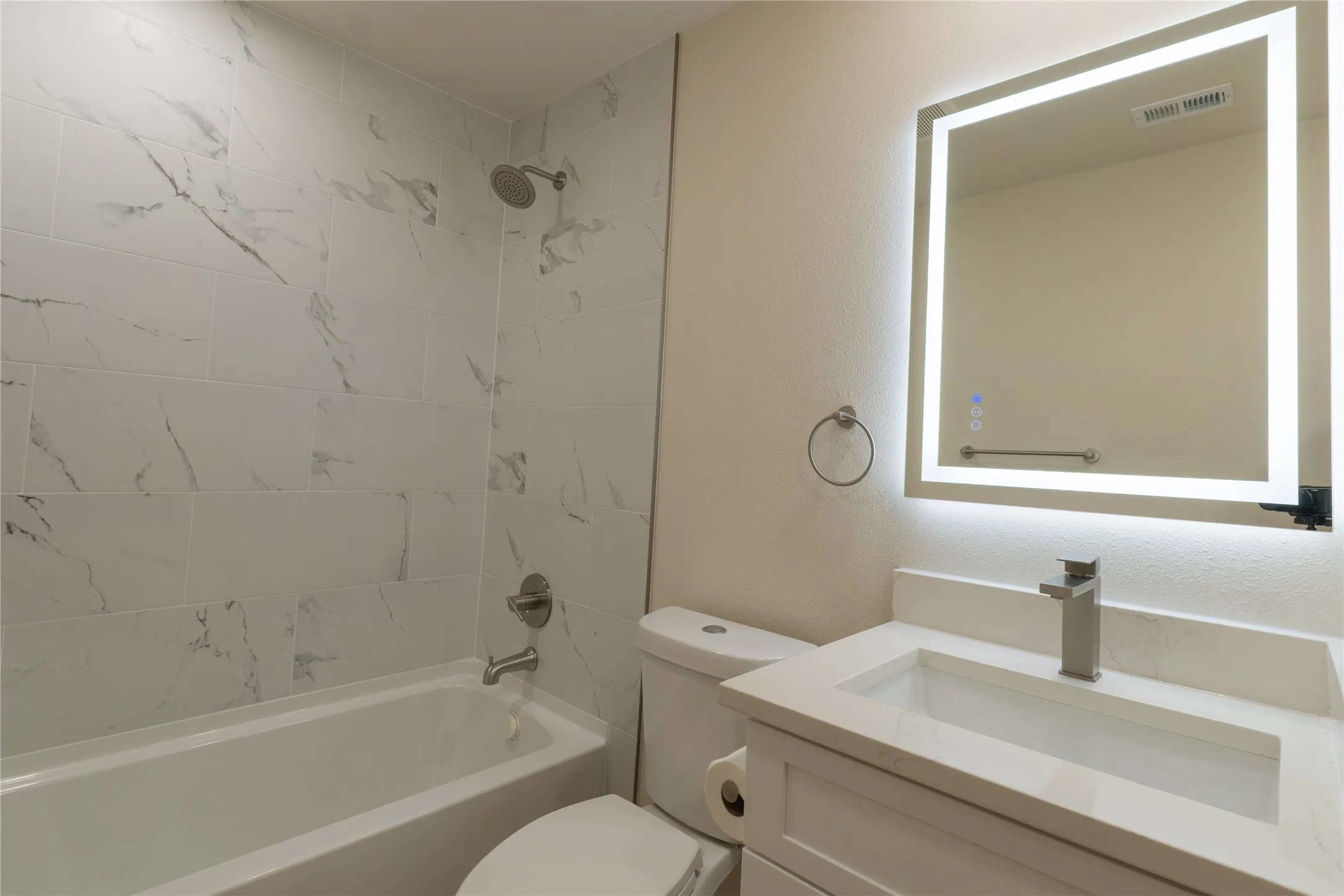 Bathroom featuring a shower-over-tub combination with large format tile surround, a vanity with a rectangular sink, brushed nickel faucet, and a back-lit mirror