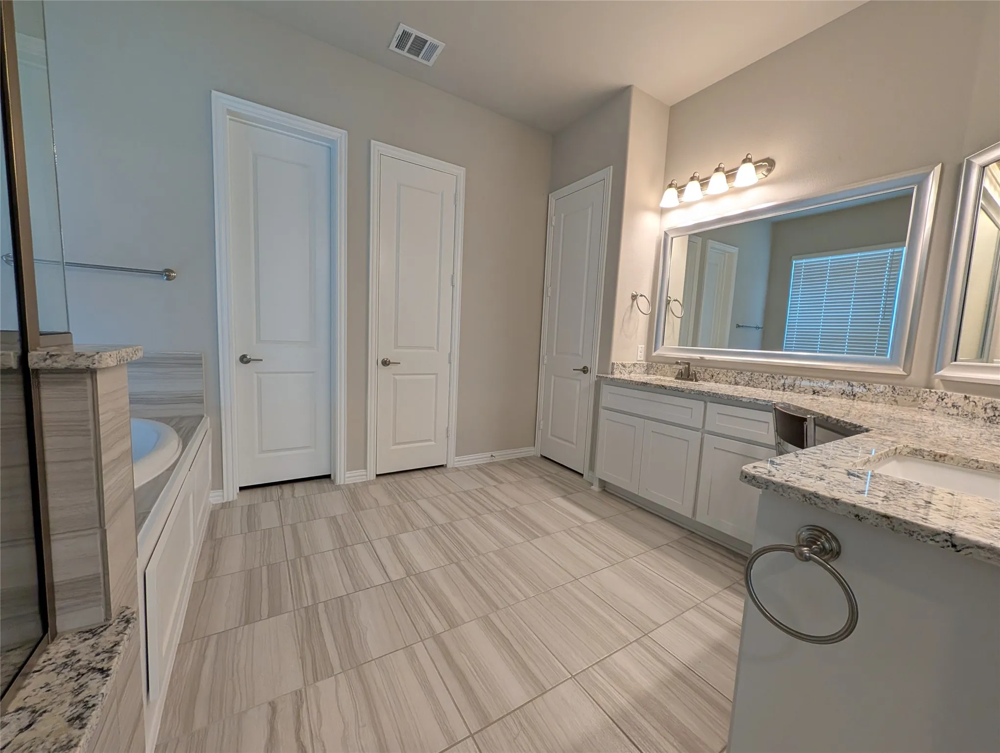 Spacious bathroom featuring light-toned tile flooring, dual vanities with granite countertops, white cabinetry, and an integrated soaking tub
