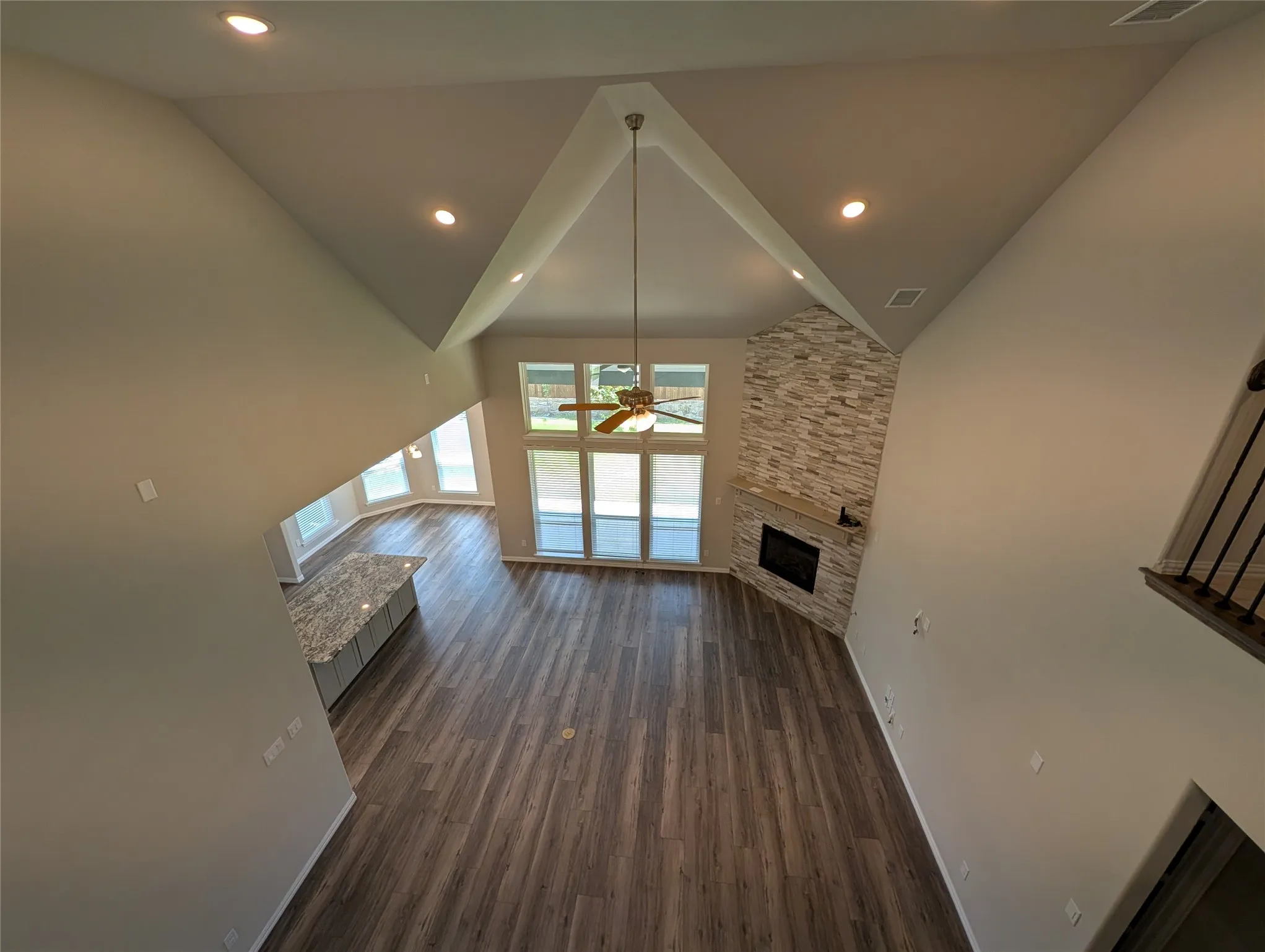 Vaulted ceiling with recessed lighting, wood-finish flooring, full-height stacked stone fireplace, large window wall, and kitchen island with granite countertop