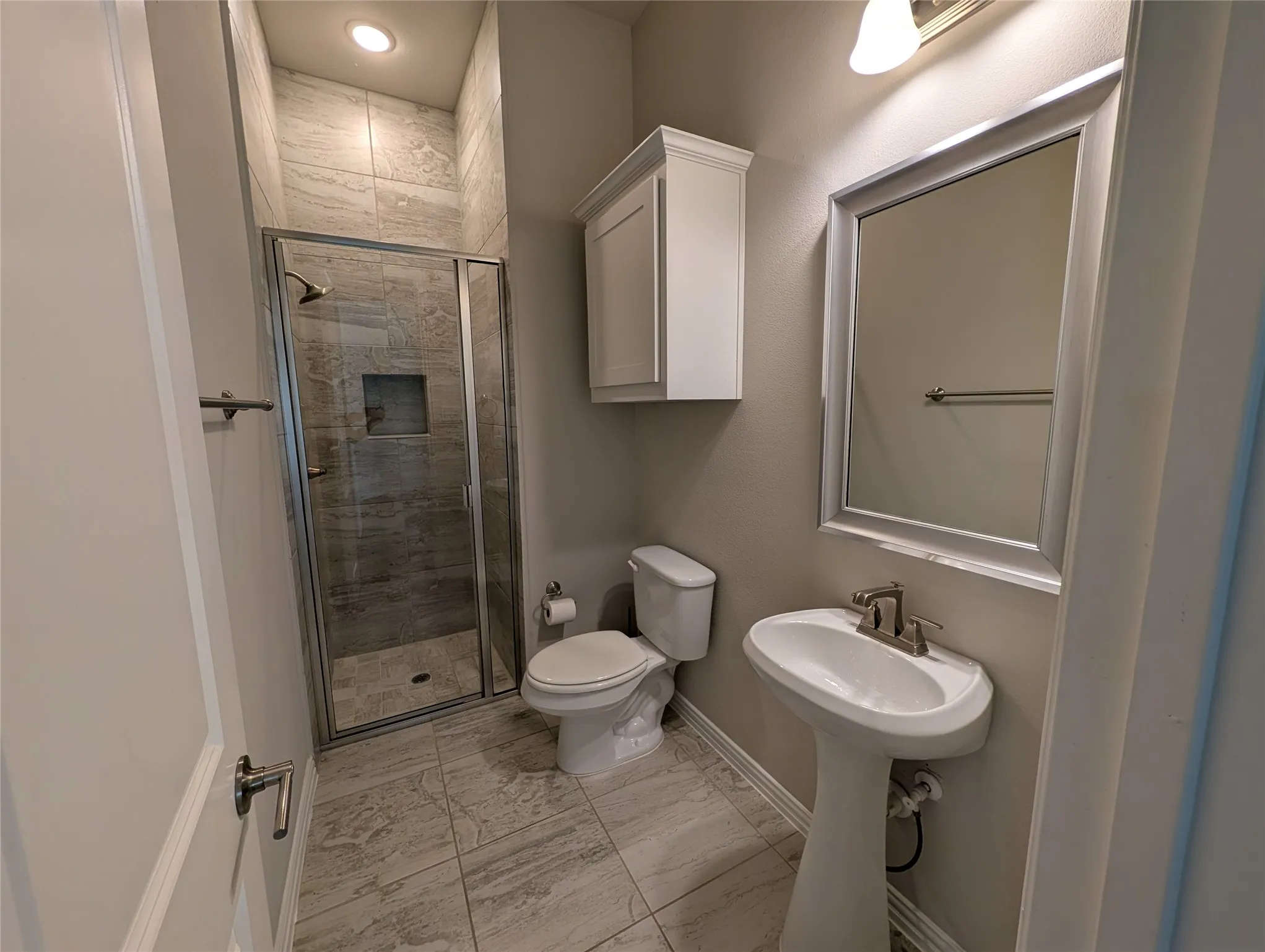 Glass-enclosed shower with a recessed shelf, pedestal sink, wall-mounted storage cabinet, and tile flooring