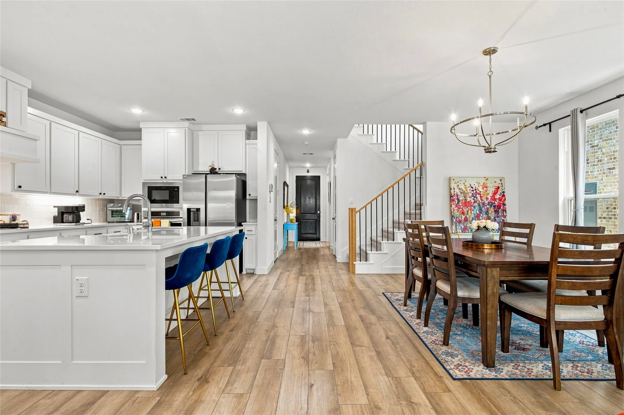 Open-concept living space featuring wood-finish flooring throughout, a modern kitchen with white cabinetry and stainless steel appliances, a dining area with a contemporary chandelier, a staircase with black metal balusters, and recessed lighting