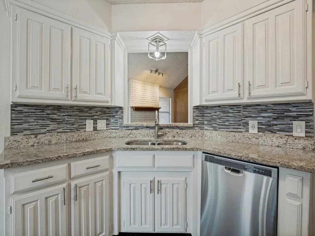 White cabinetry with paneling, granite-style countertops, mosaic tile backsplash, stainless steel dishwasher, and a double basin sink