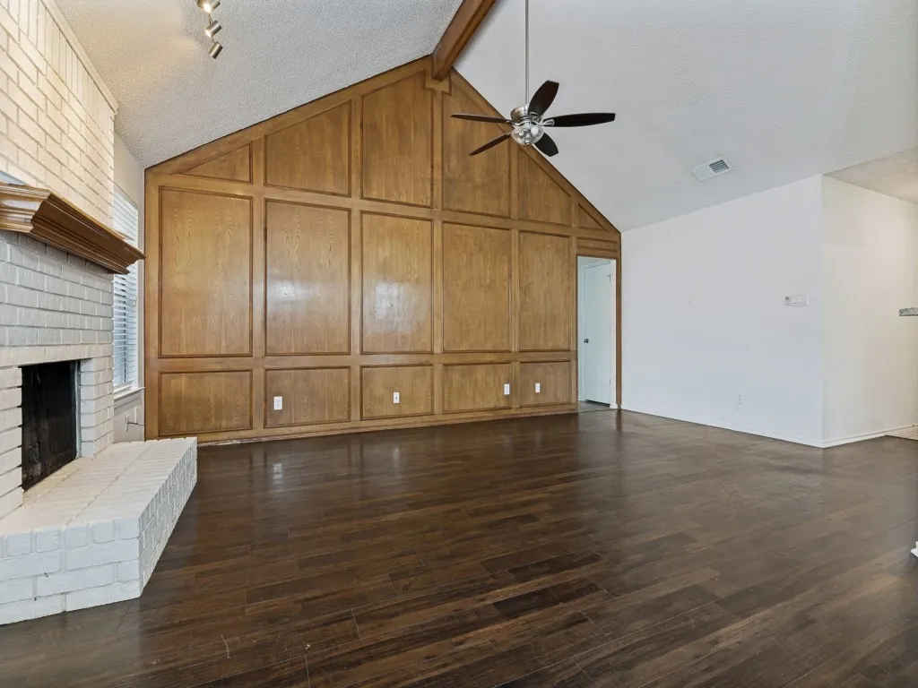 Vaulted ceiling with exposed wood beam, wood-finish flooring, paneled accent wall, painted brick fireplace with hearth, and ceiling fan