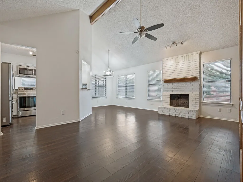 Vaulted ceiling with exposed wood beam, wood-finish flooring, white brick fireplace with wood mantel, ceiling fan, and stainless steel appliances in the adjacent kitchen