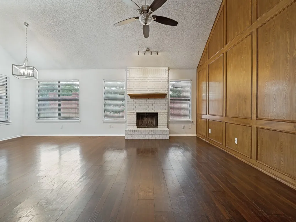 Vaulted ceiling with a ceiling fan, wood-finish flooring, white brick fireplace, multiple windows with blinds, and a wood-paneled accent wall