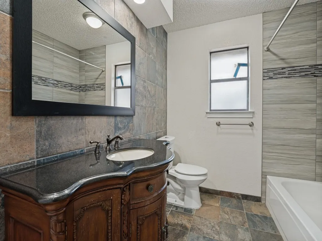 Custom vanity with an undermount sink and dark countertop, framed mirror, tiled accent wall, tiled shower surround with a decorative band, and tiled flooring