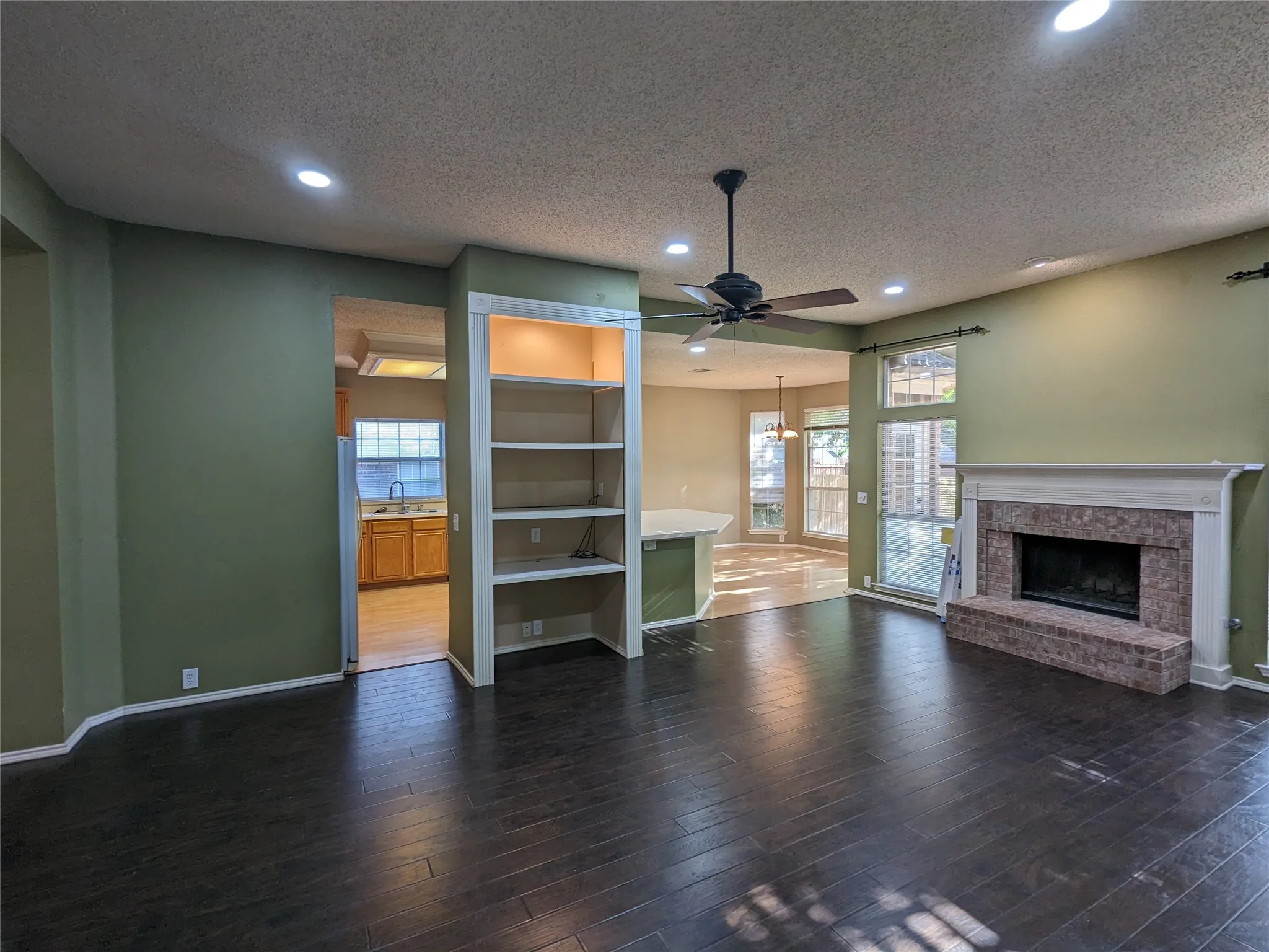 Wood-finish flooring, built-in shelving, ceiling fan, fireplace with a brick surround and white mantel, and recessed lighting