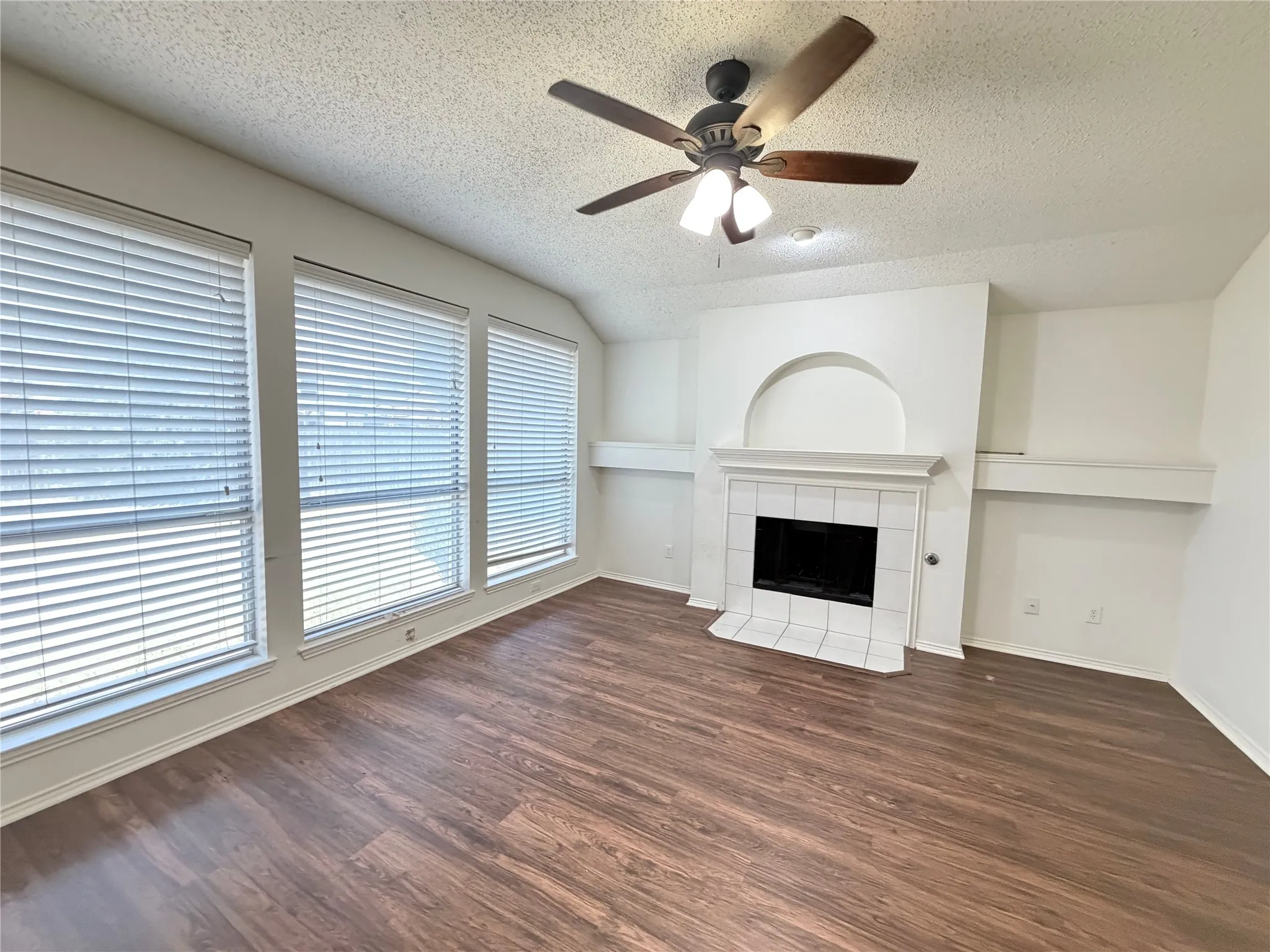 Wood-finish flooring, three large windows with blinds, a ceiling fan with integrated lighting, a fireplace with a white tile surround, and built-in shelving