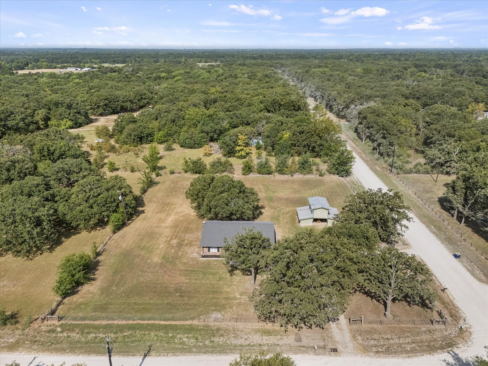 Expansive wooded surroundings with a gravel road, two distinct structures, and a clear sky overhead