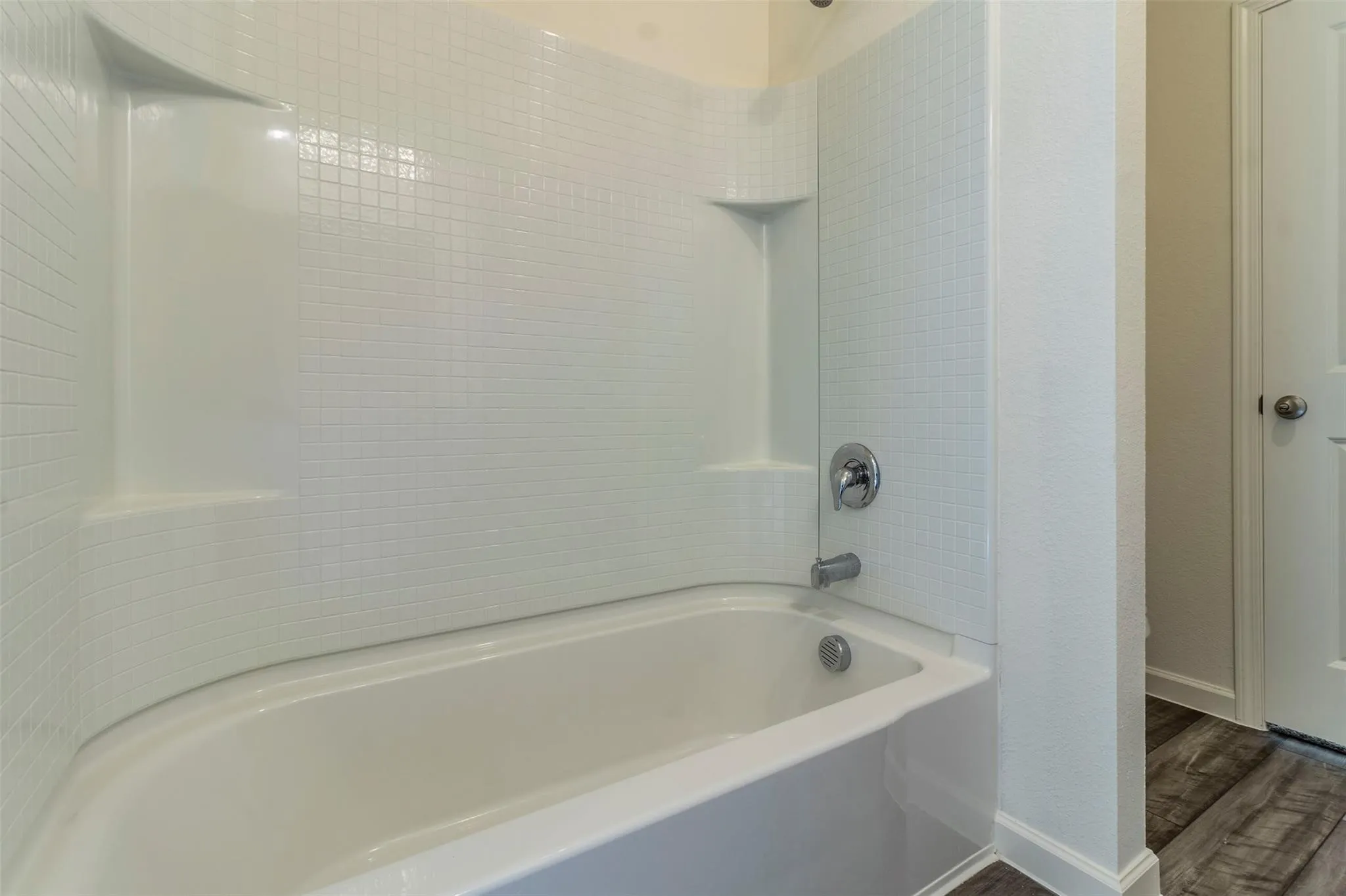 White tub and shower combination with a textured tile-patterned surround, integrated shelving, and chrome fixtures