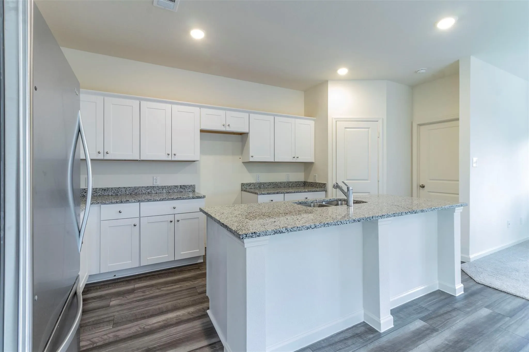 Kitchen featuring white shaker-style cabinetry, granite countertops, a center island with an undermount sink, stainless steel refrigerator, and wood-finish flooring