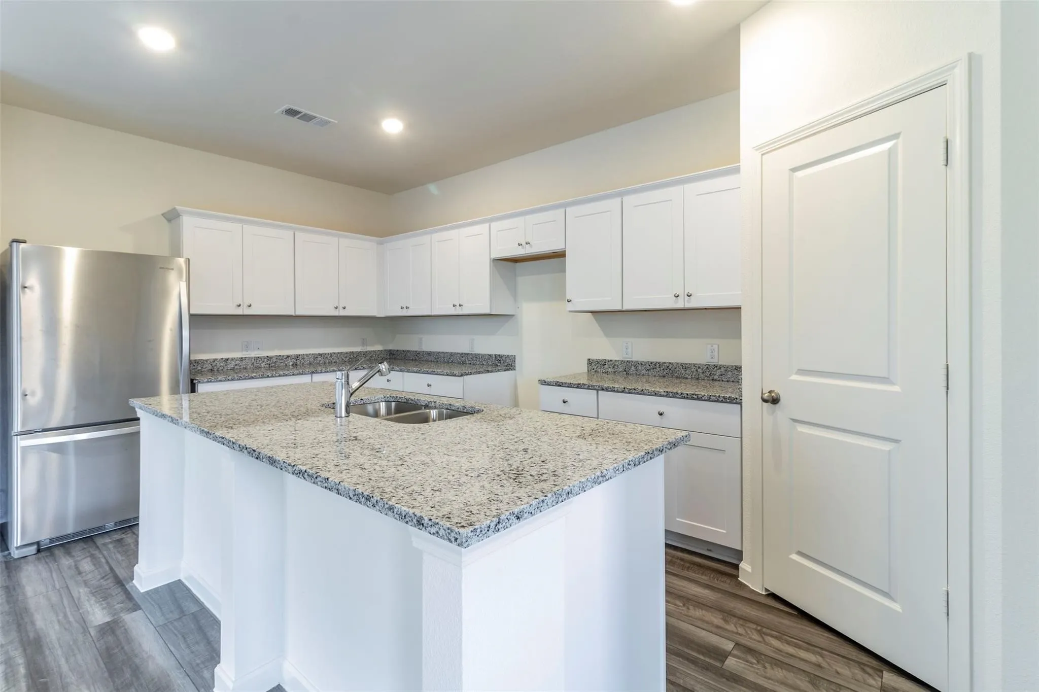 Kitchen featuring white cabinetry, light-toned countertops, a stainless steel refrigerator, a kitchen island with an integrated sink, and wood-finish flooring
