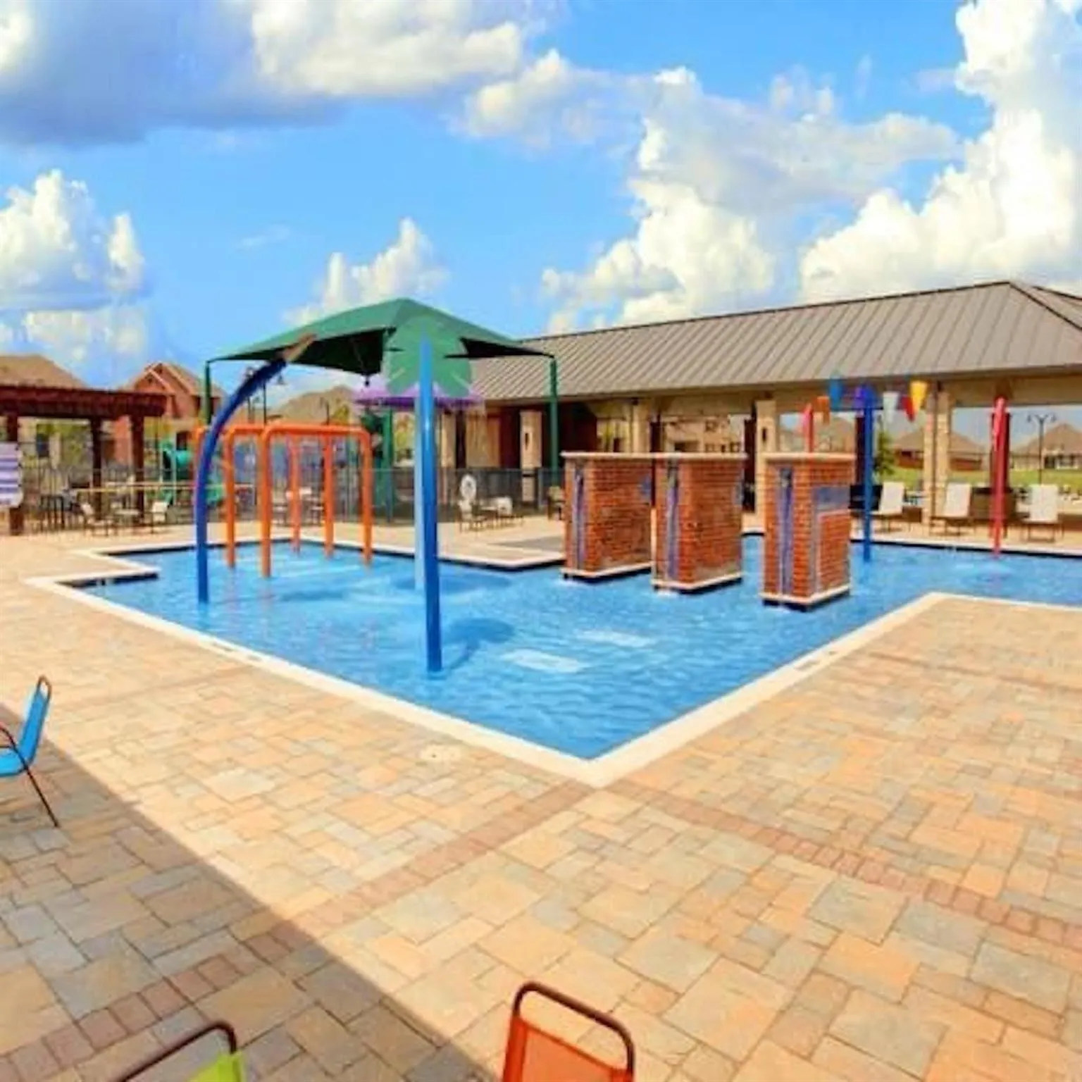 Water feature with multiple spray elements, surrounding paver decking, and an adjacent covered outdoor gathering area