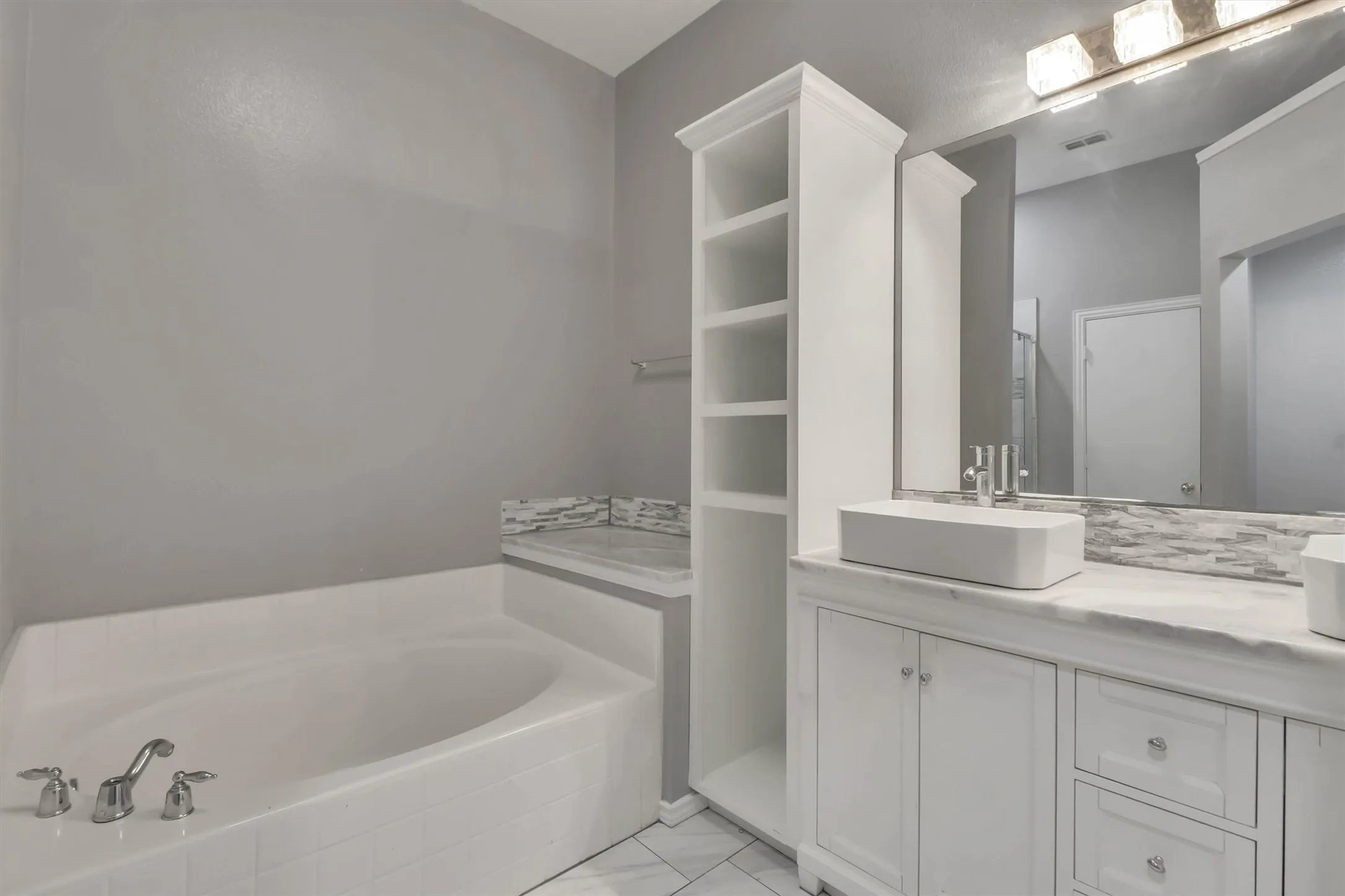 Built-in garden tub, white vanity with integrated sink, stone countertop, tile flooring, and built-in shelving