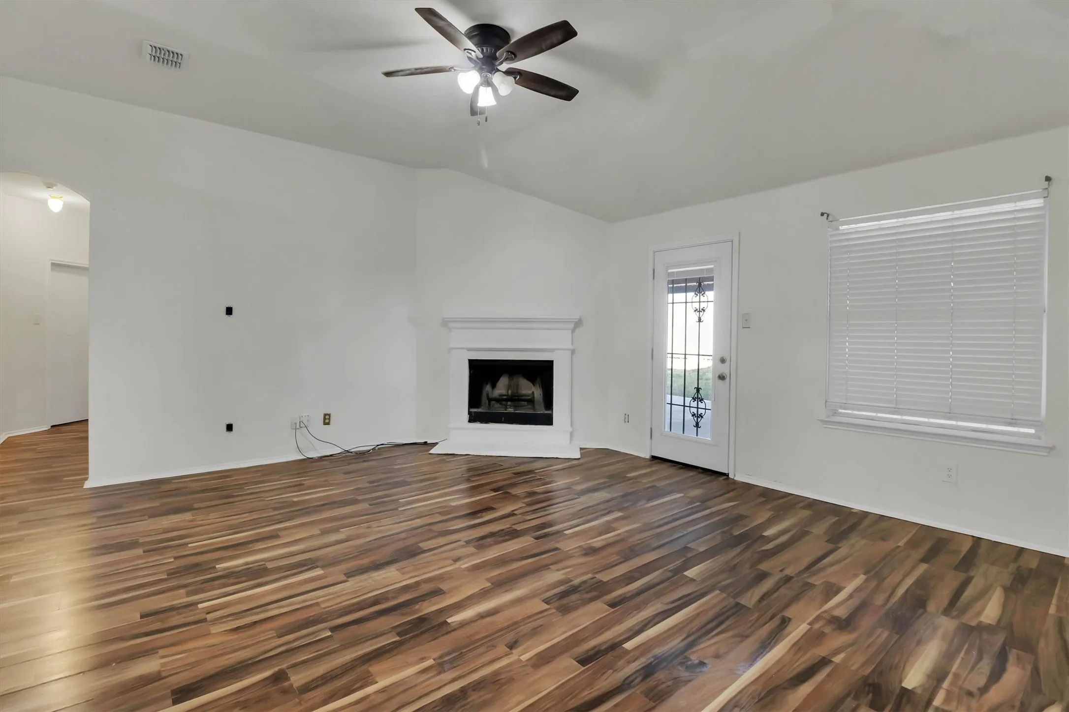 Wood-finish flooring, vaulted ceiling, corner fireplace with white mantel, ceiling fan, and window with blinds