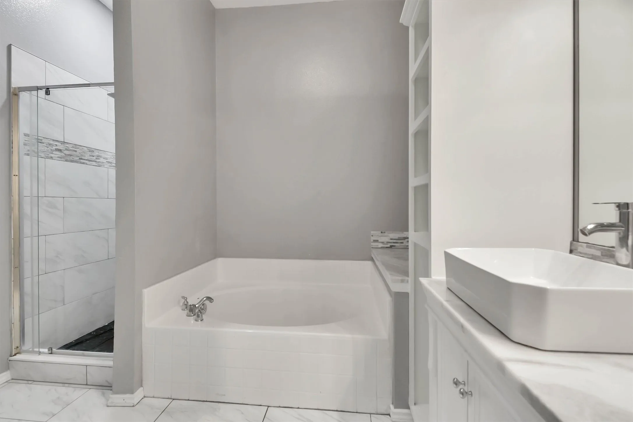Built-in soaking tub, glass-enclosed shower with tiled surround, white vanity with vessel sink, polished chrome fixtures, and built-in shelving