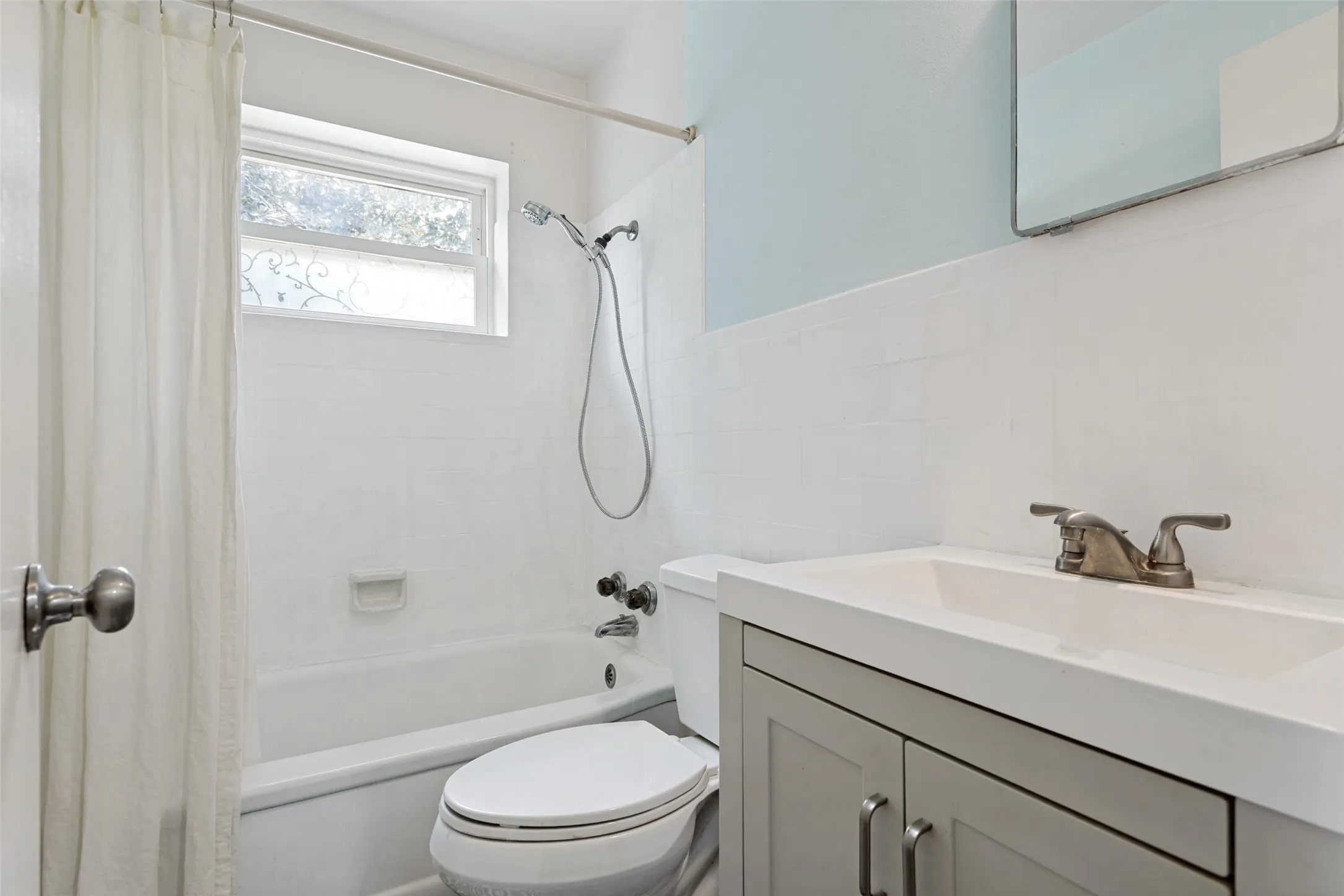 Bathroom featuring a white bathtub, tiled shower surround, window with privacy glass, single-basin vanity with gray cabinetry, and a toilet