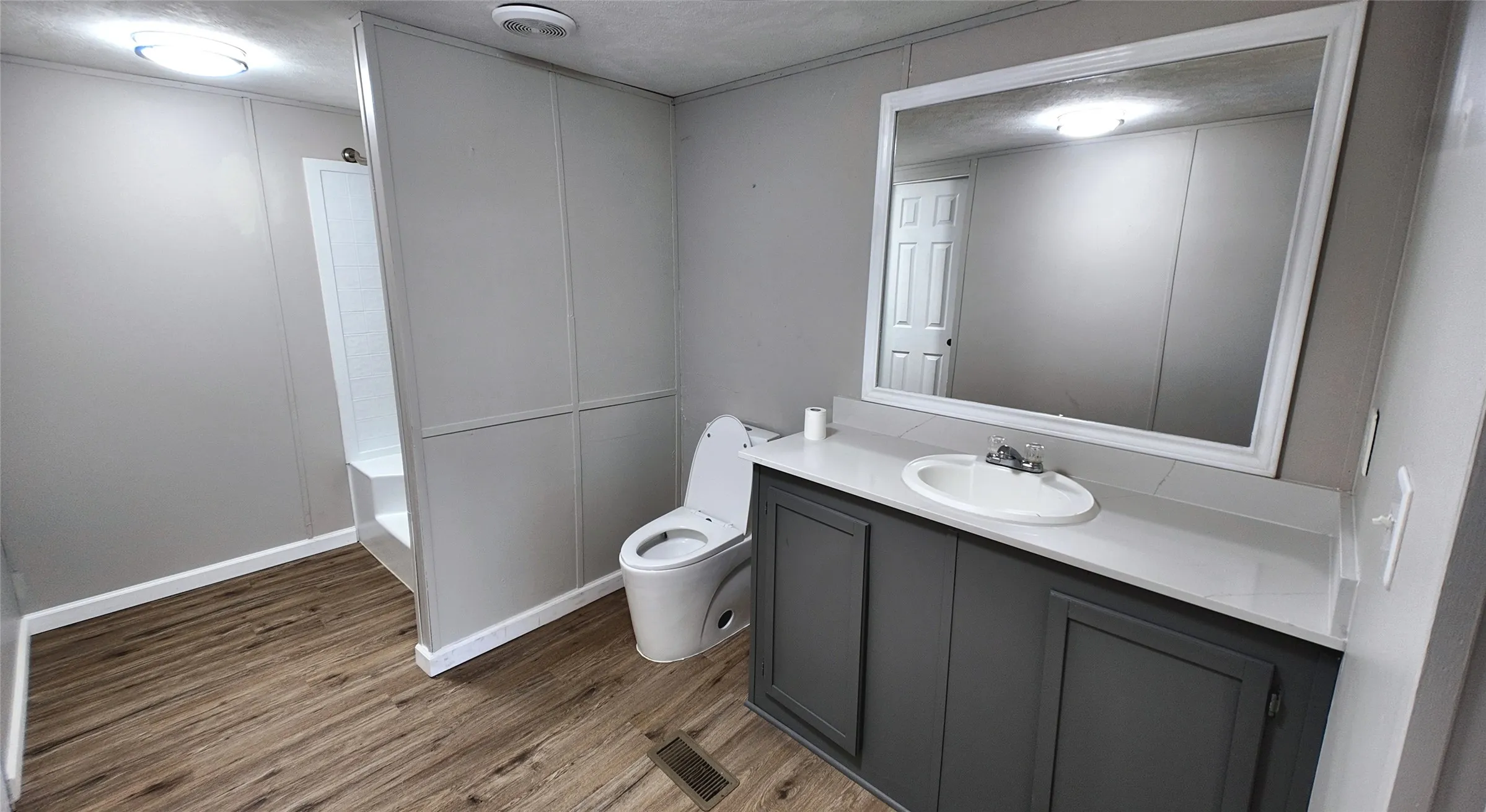 Bathroom featuring wood-finish flooring, a single vanity with a white countertop and dark gray cabinetry, a framed mirror, and a built-in shower