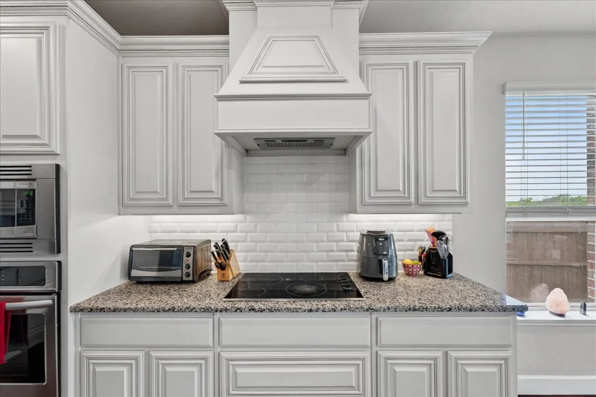 Kitchen featuring white shaker-style cabinetry, a built-in range hood, subway tile backsplash, speckled countertops, and an electric cooktop