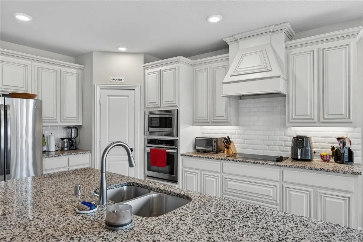 Spacious kitchen featuring an expansive granite island with a double basin sink, white shaker-style cabinetry, subway tile backsplash, and stainless steel appliances, including a built-in double oven