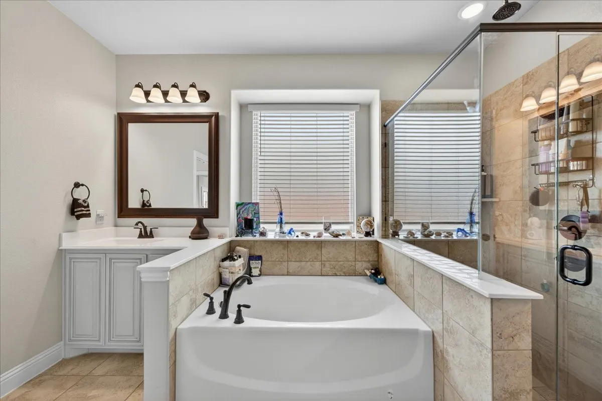 Full bathroom featuring a built-in soaking tub with dark bronze fixtures, a tiled walk-in shower with glass enclosure, a single vanity with white cabinetry and countertop, and a window with blinds