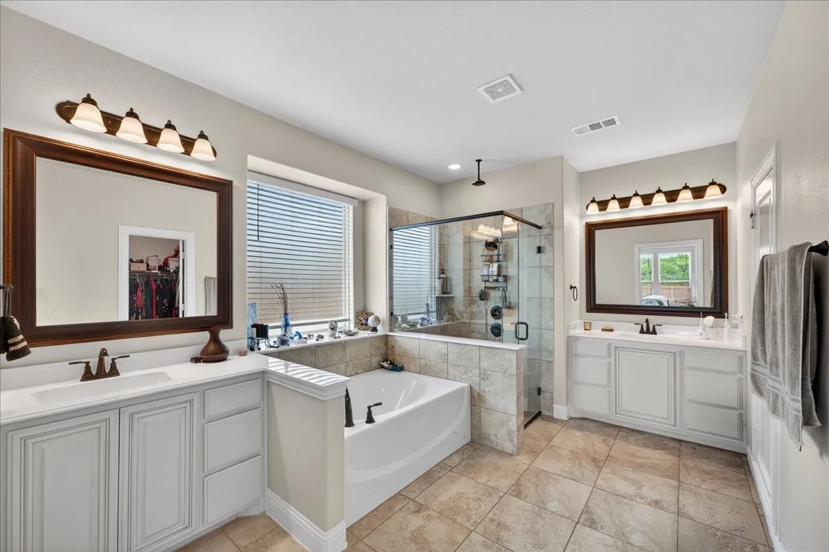 Bathroom featuring a soaking tub, glass-enclosed shower, dual vanities with white cabinetry, large framed mirrors, and tile flooring