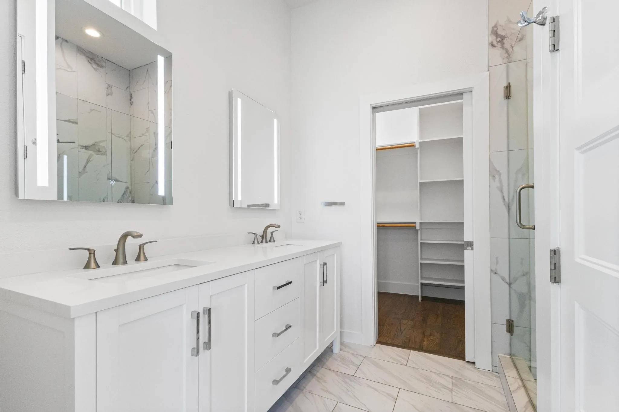 Dual white vanities with integrated sinks, illuminated mirrors, tiled flooring, and a walk-in closet with built-in shelving
