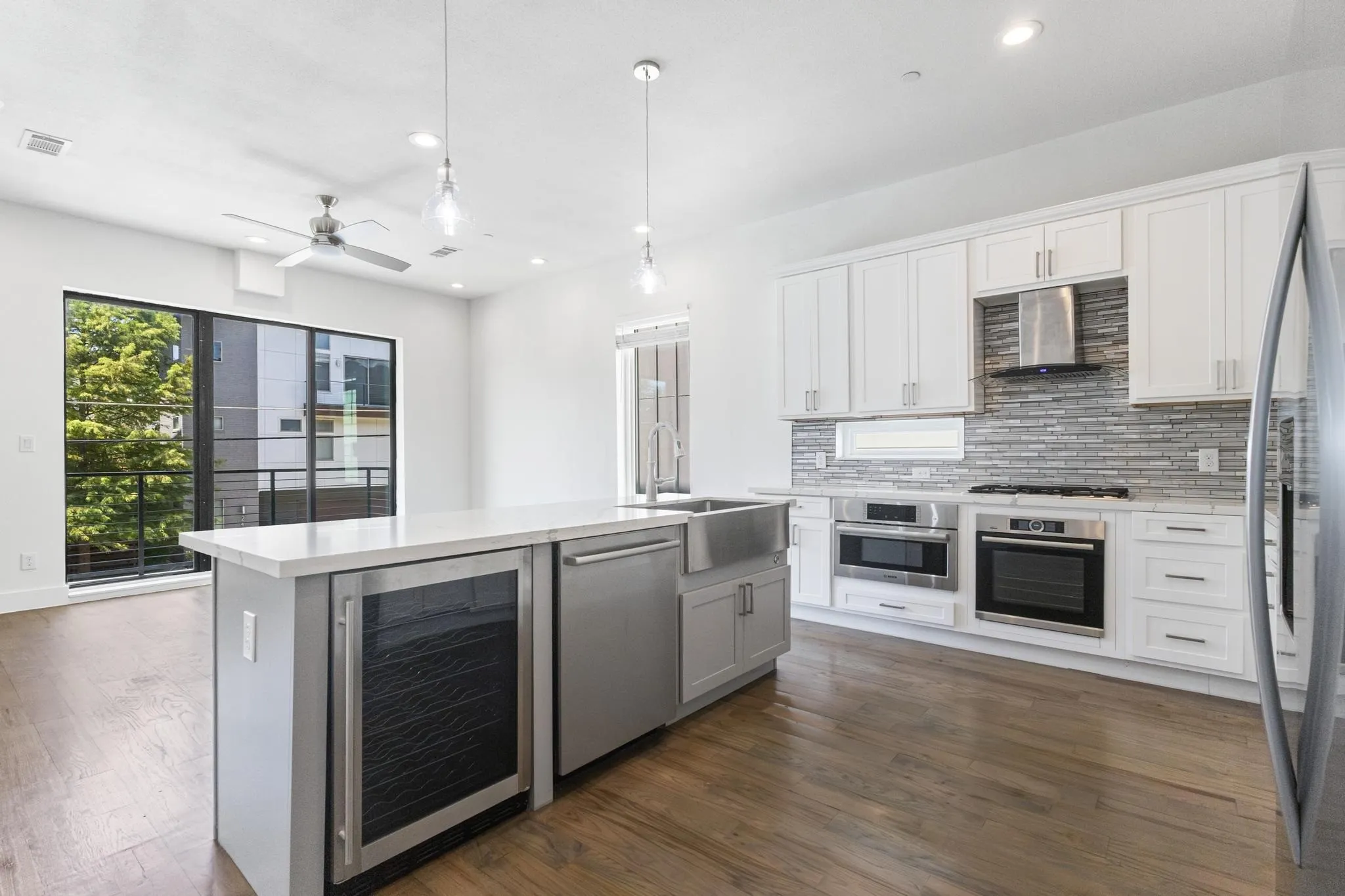 Open-concept kitchen with wood-finish flooring, white cabinetry, stainless steel appliances, and a central island featuring a beverage cooler and farm sink