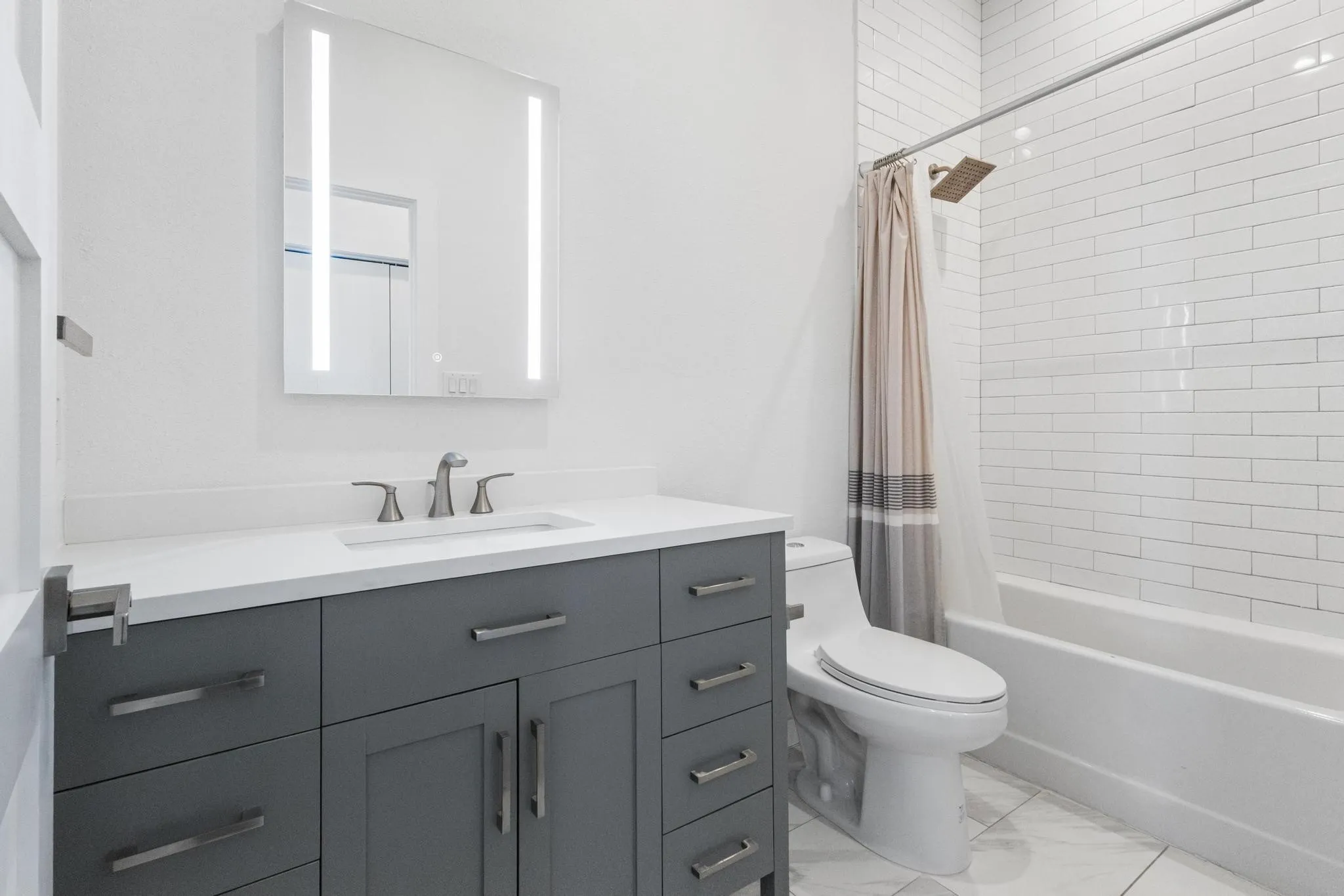 White subway tile tub surround, sleek vanity with a quartz countertop, integrated rectangular sink, illuminated mirrored medicine cabinet, and contemporary floor tiling