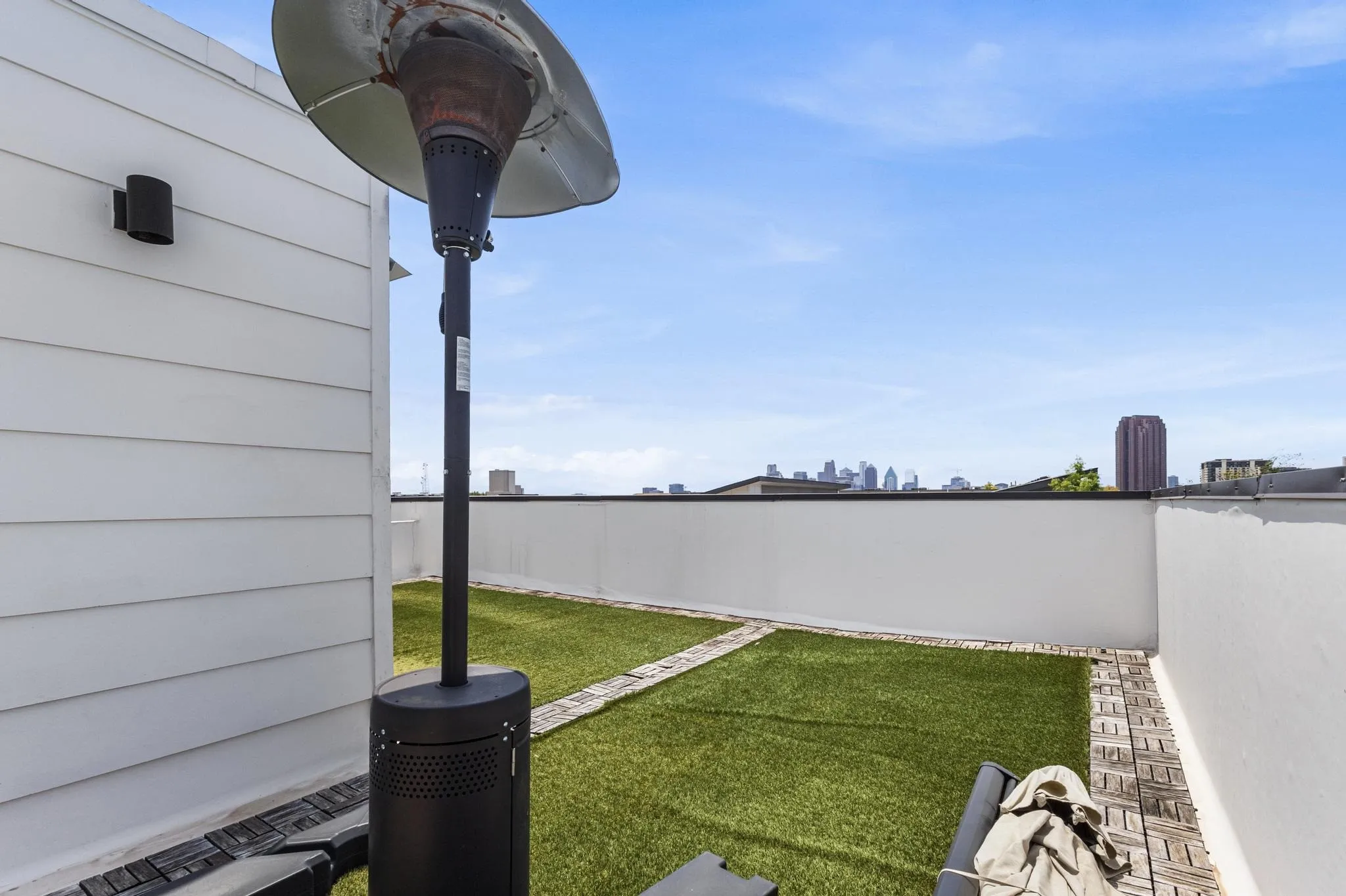 Rooftop terrace featuring white siding, a permanent outdoor light fixture, artificial turf, and a city skyline in the distance