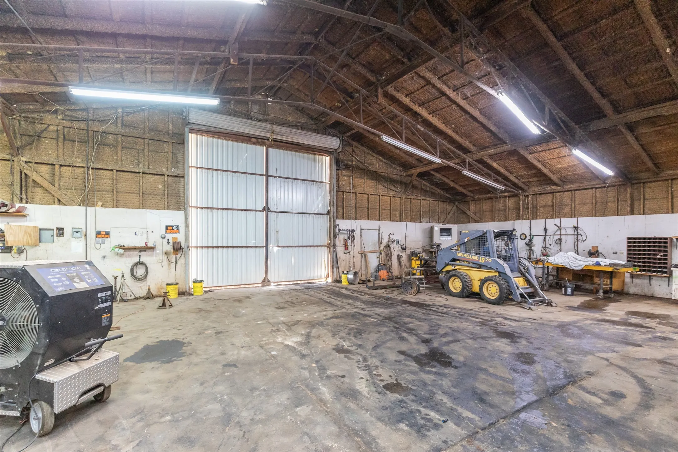 Spacious interior work area featuring a high vaulted ceiling with exposed wood beams, concrete flooring, and multiple fluorescent light fixtures