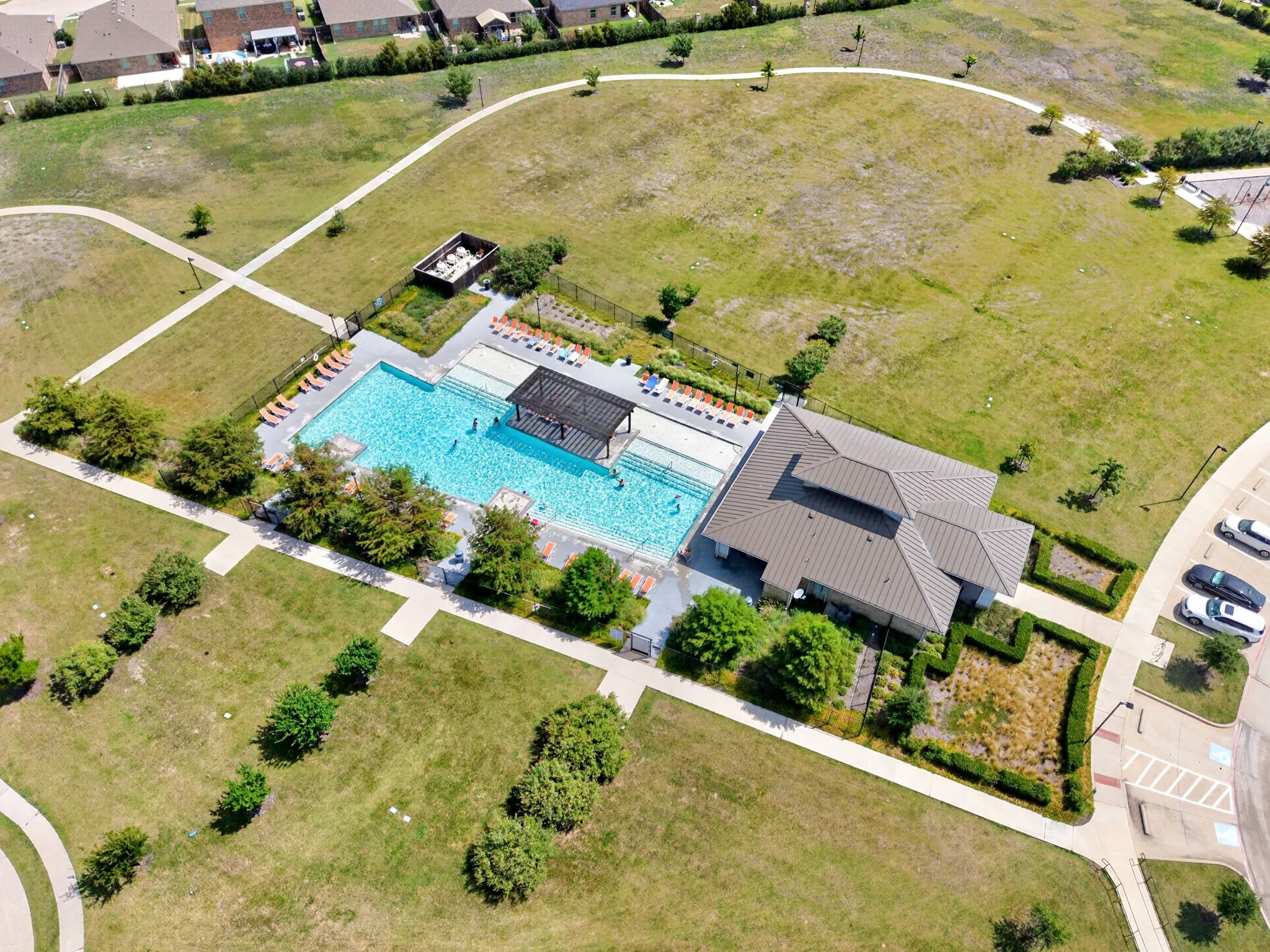 Community pool with a shaded pergola, adjacent building with a multi-level roof, paved pathways, open grassy areas, and dedicated parking